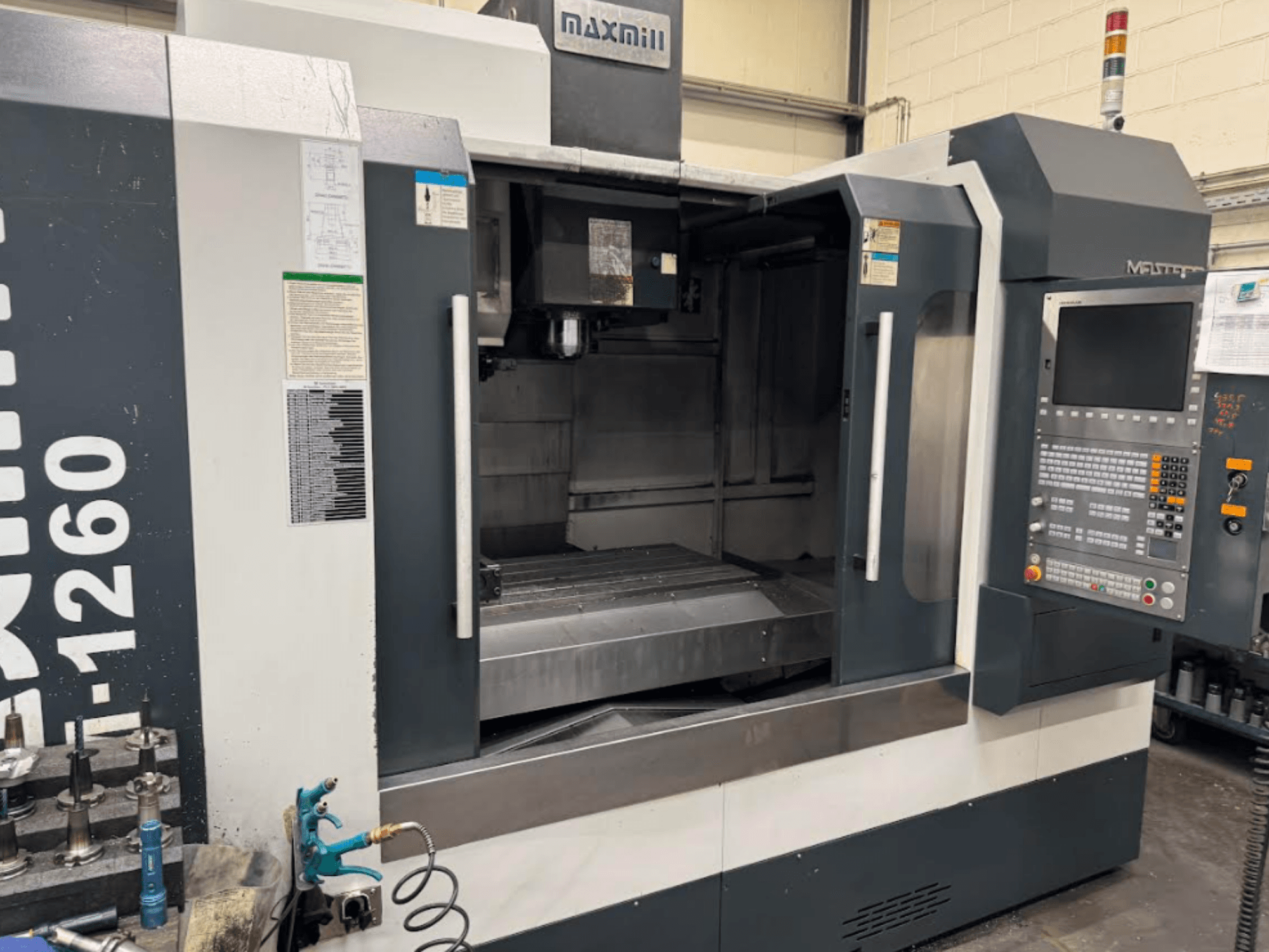 Fresatrice CNC Maxmill, modello M-1260, vista frontale che mostra l'area di lavoro vuota con il pannello di controllo e le istruzioni operative.