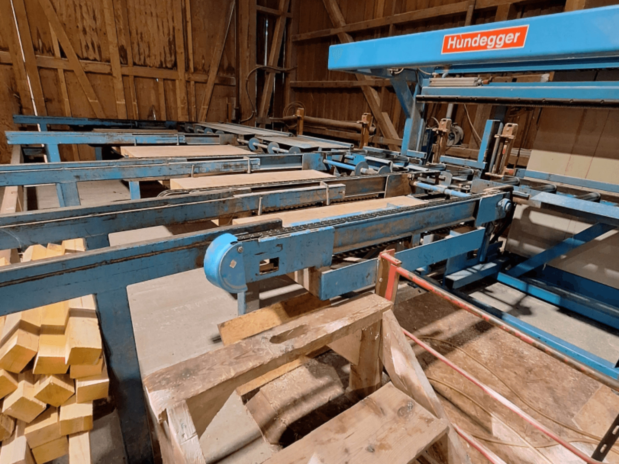 Centro di lavorazione del legno a controllo numerico HUNDEGGER K2-A all’interno di un fienile, caratterizzato da una struttura metallica blu, un sistema di trasporto e travi di legno impilate.