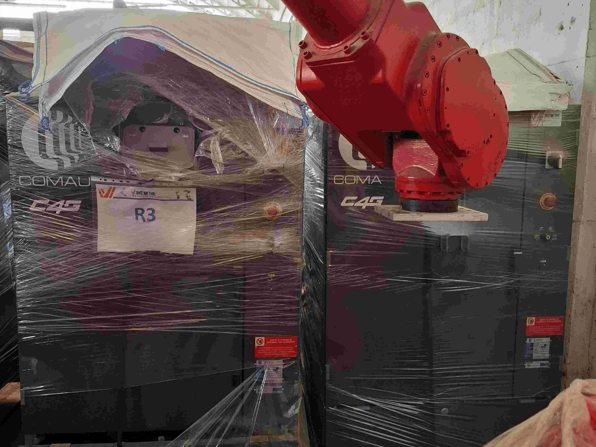 Braccio robotico Comau C4G visto dal davanti, avvolto nella plastica, etichettato "R3", con adesivi di attenzione visibili sulla macchina.