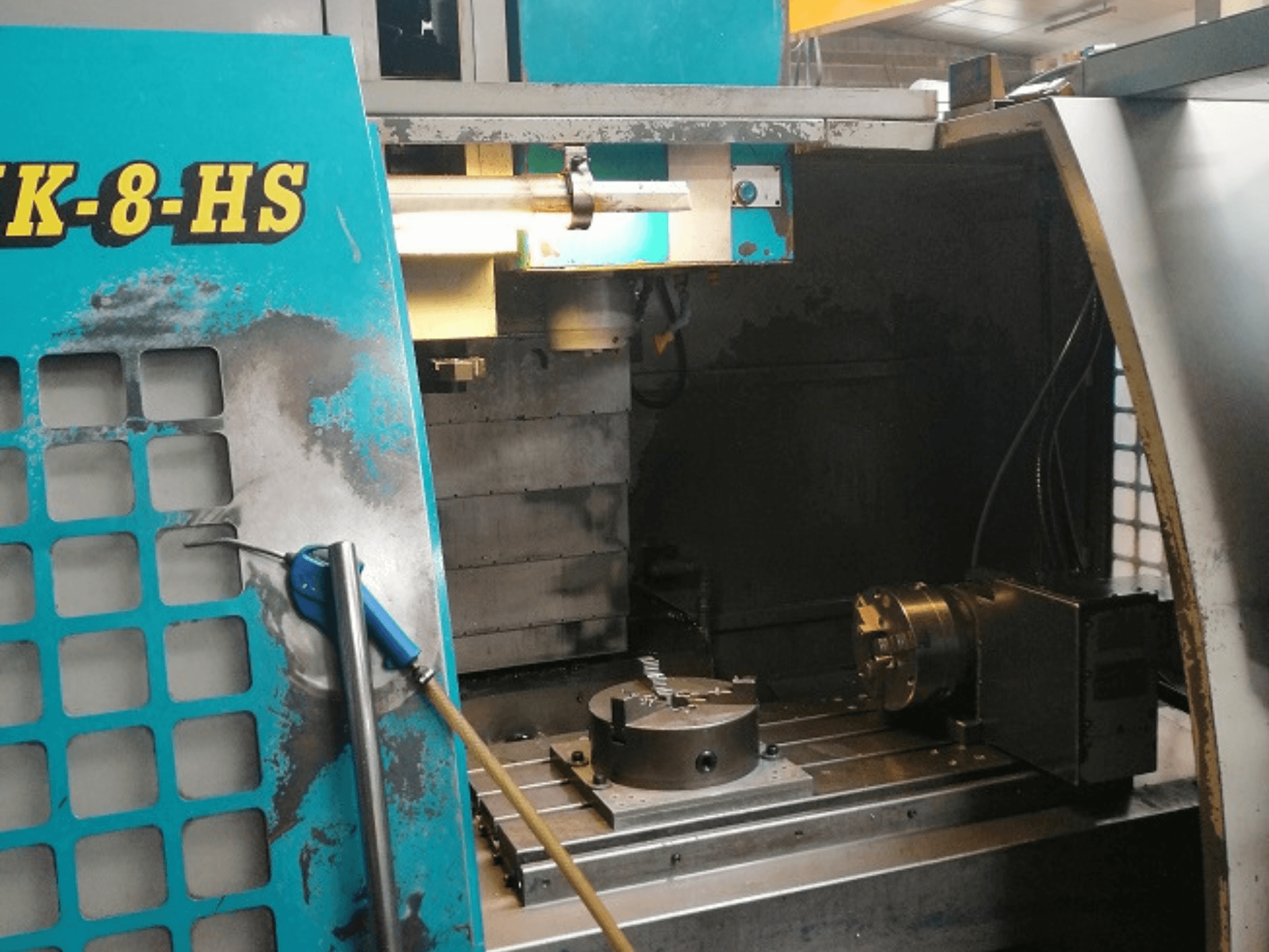 Vista frontale di una macchina CNC SK-8-HS blu, che mostra un piano di lavoro con morsa e pannello di controllo; segni di usura e di grasso visibili.