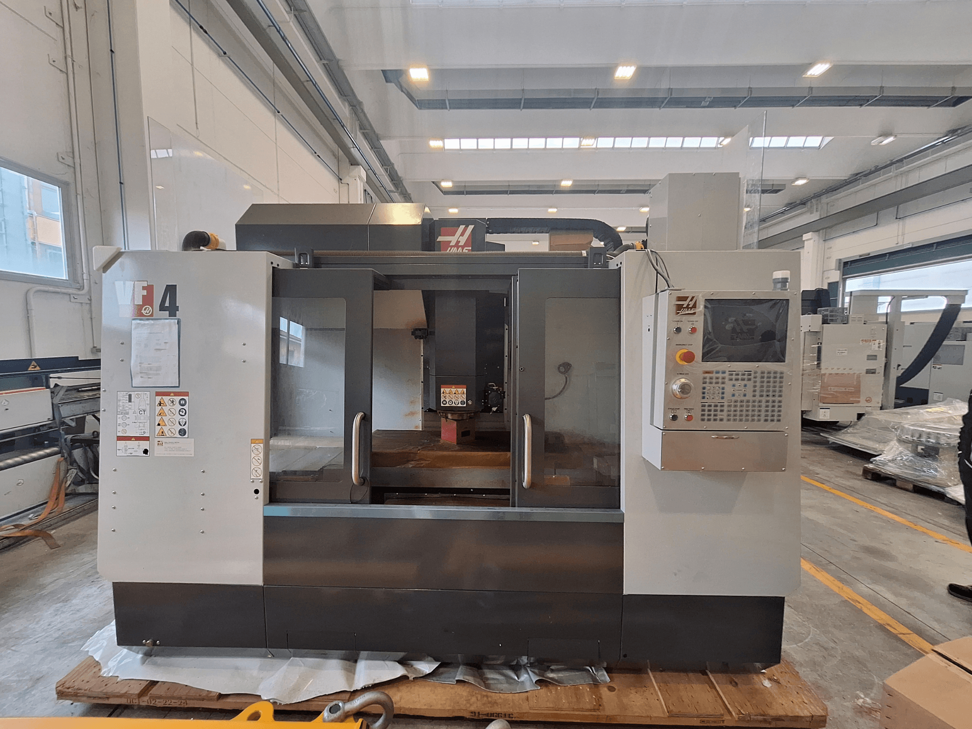 Centro di lavorazione verticale HAAS VF-4, vista frontale, che mostra il pannello di controllo, l'area di lavoro e le etichette di sicurezza in un ambiente di magazzino.
