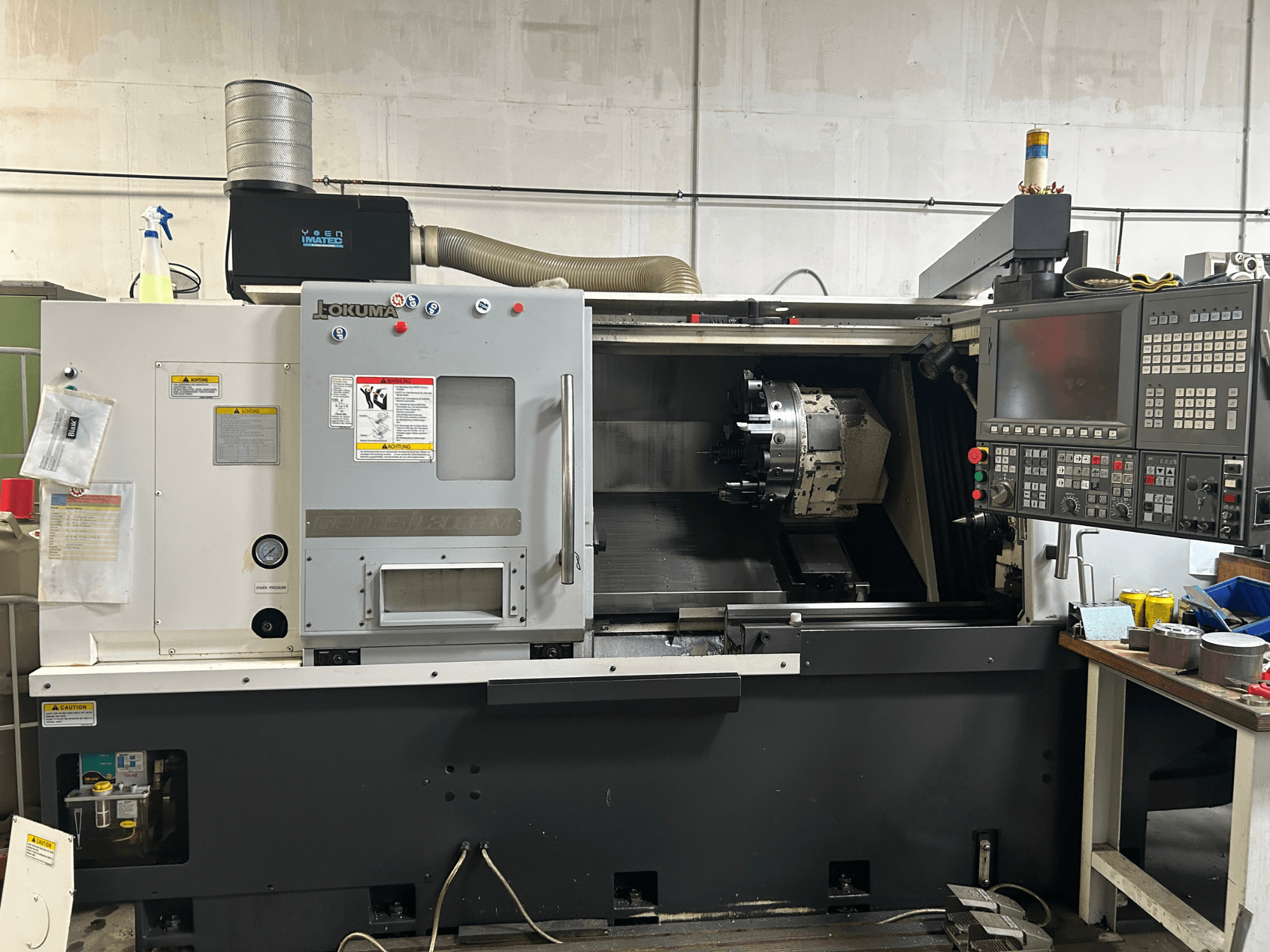 Vista frontale di un tornio orizzontale Okuma Genos L300E-M con pannello di controllo, mandrino visibile e vari strumenti di impostazione.