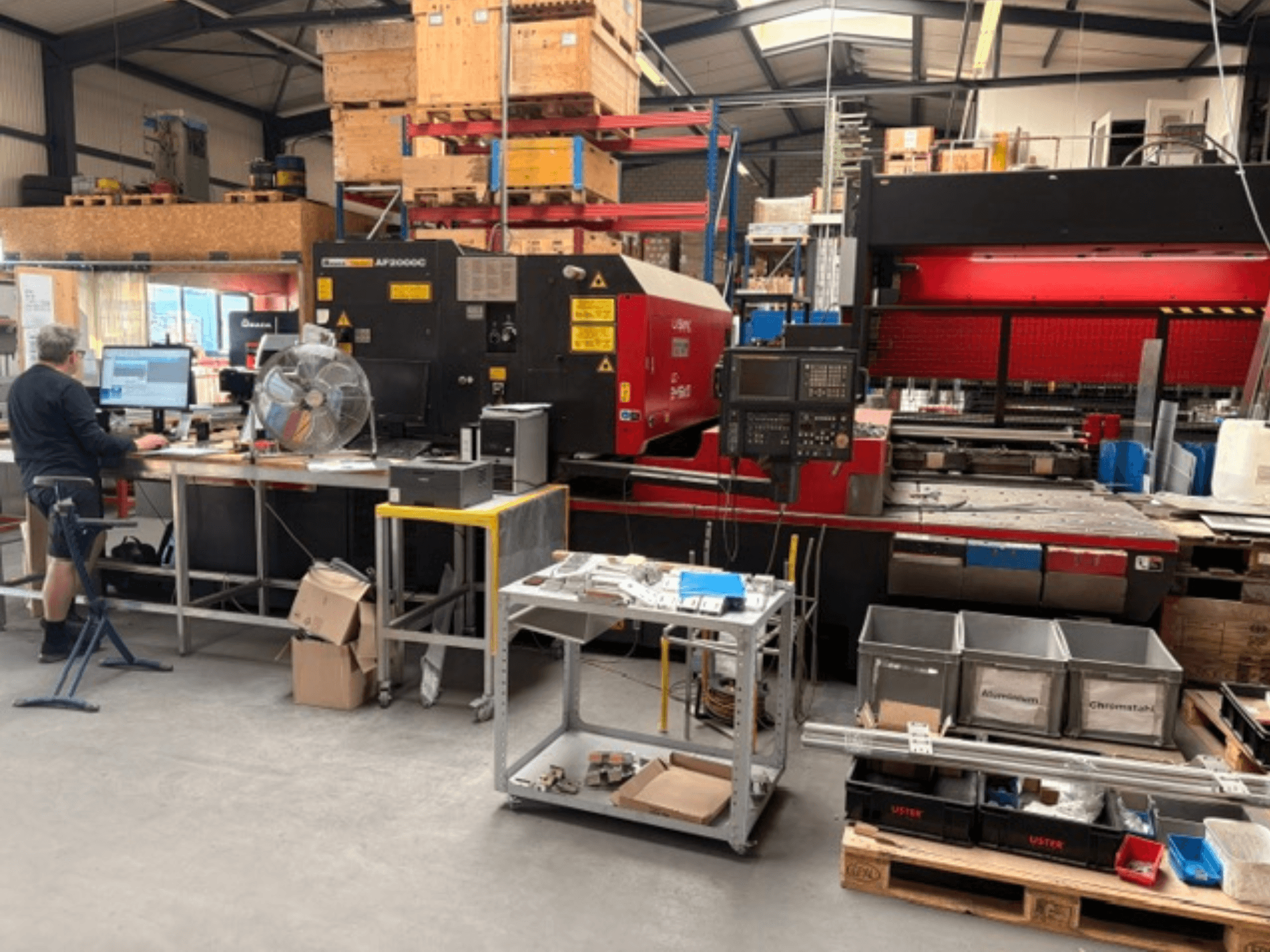 Macchina per il taglio laser a CO₂ AMADA LC-2415αIII in un ambiente di officina, con una postazione di lavoro informatica visibile in primo piano.