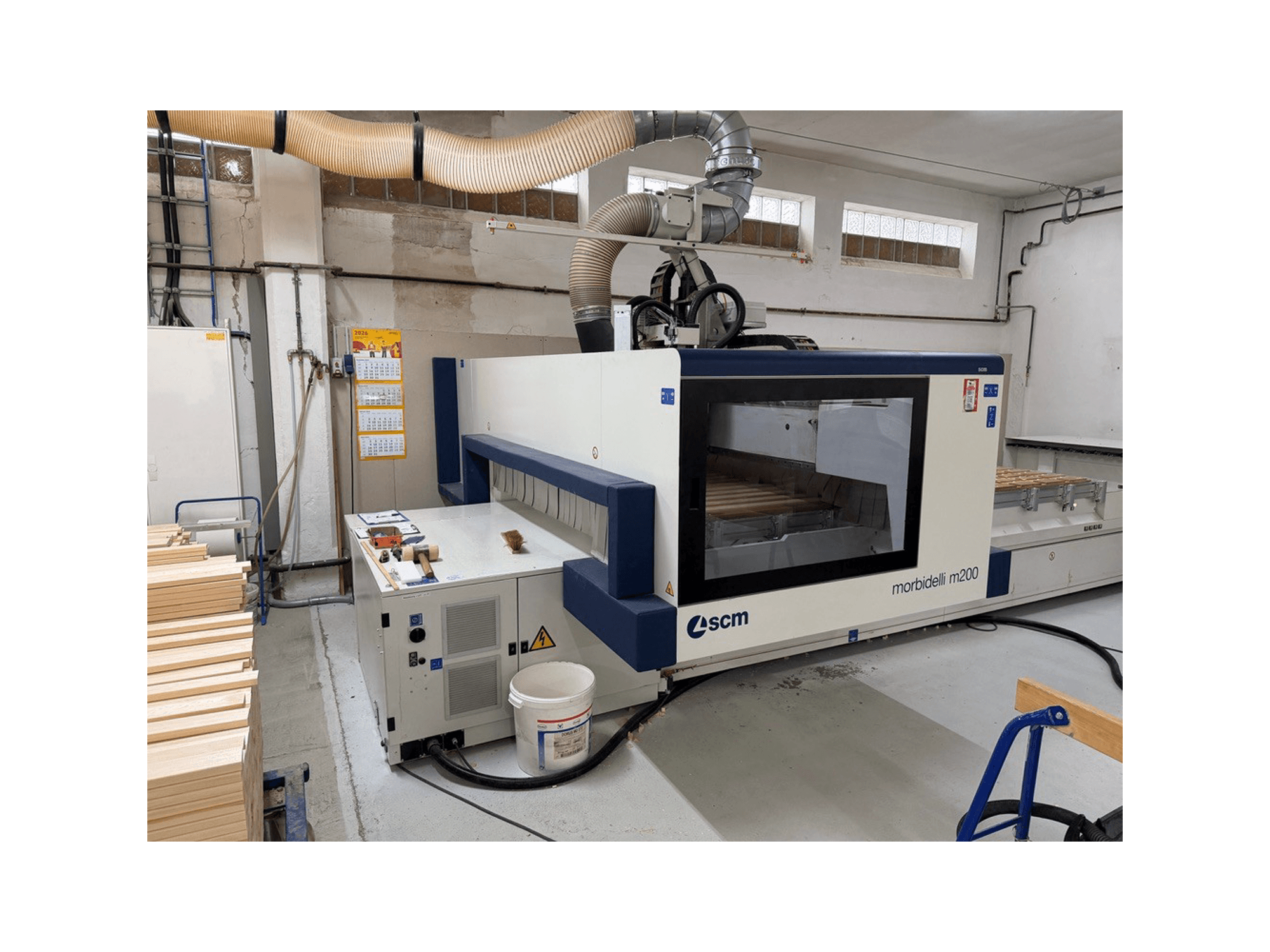 Centro di lavorazione CNC SCM Morbidelli M200F in un'officina, vista laterale con pezzi di legno sul tavolo e sistema di ventilazione sopra.