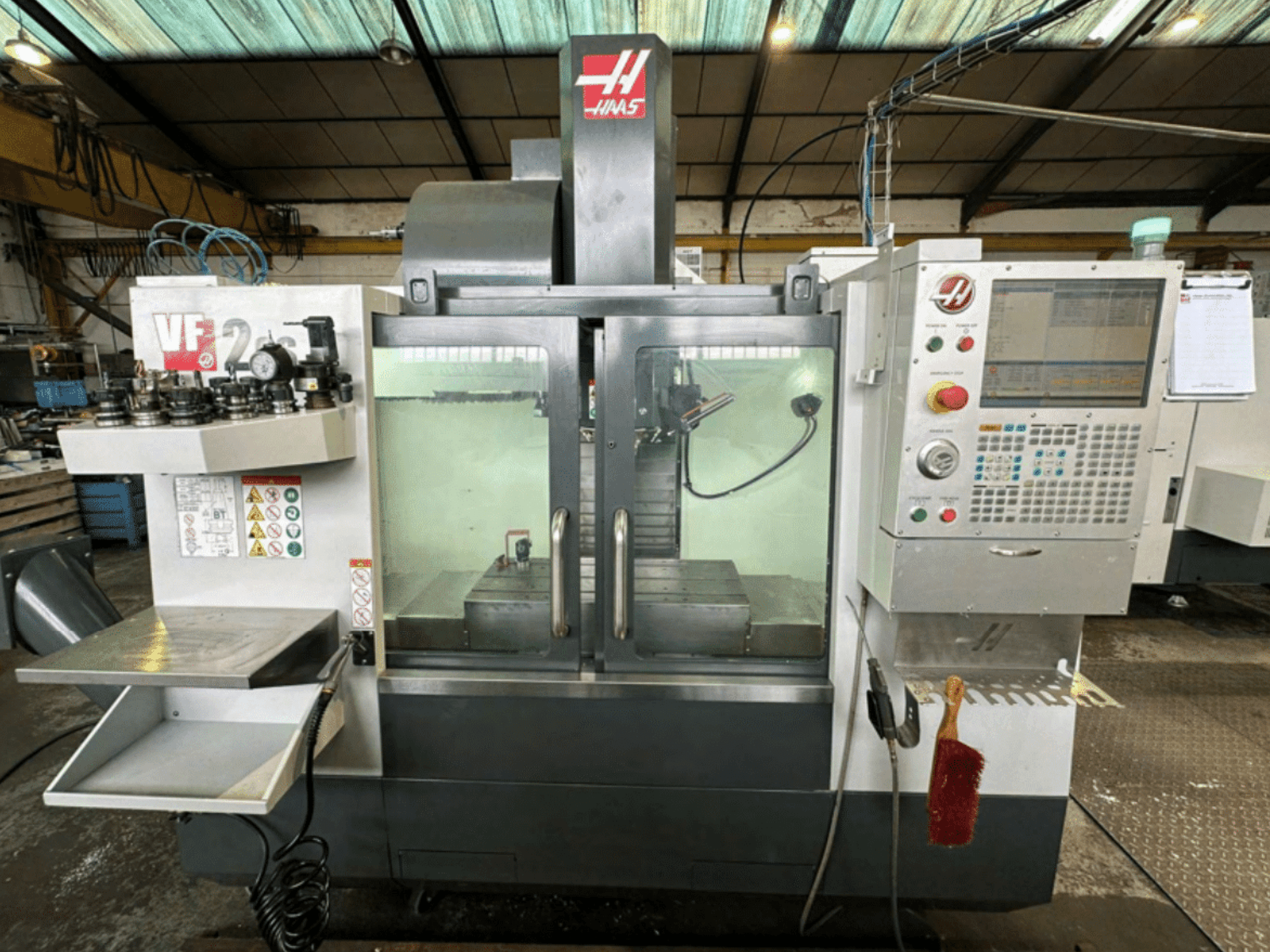 Vista frontale della macchina CNC Haas VF2, che mostra il pannello di controllo, i portautensili e l'area di lavoro con etichette di sicurezza e una spazzola.