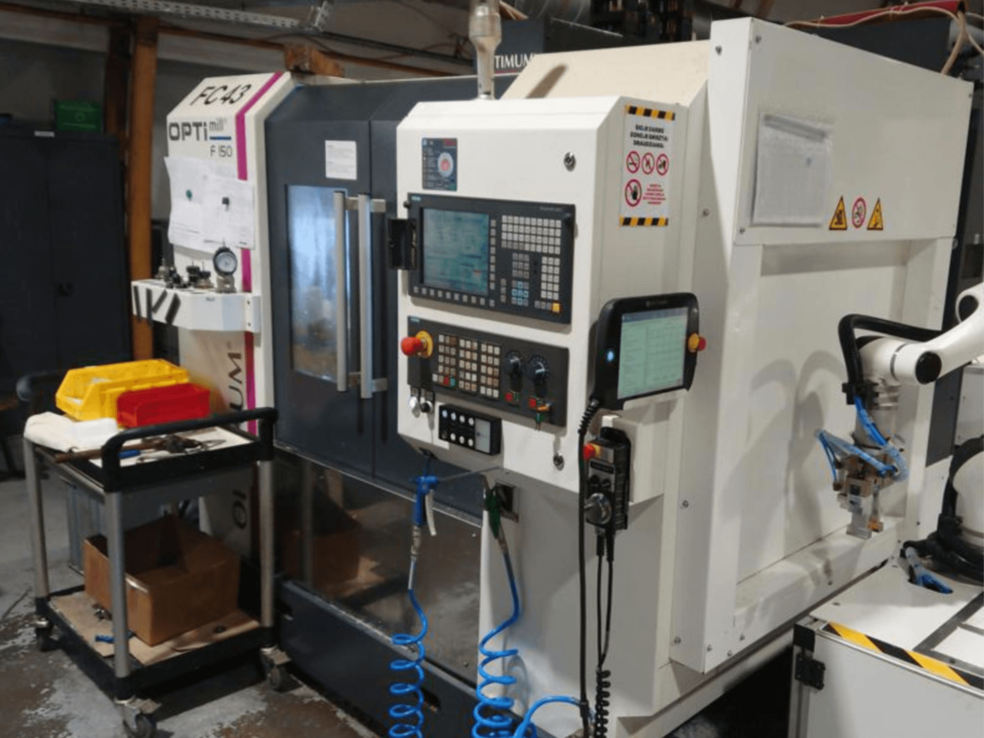 Pannello di controllo della fresatrice OPTIMUM F50 con tubi, utensili e braccio robotico visibili in un'officina.