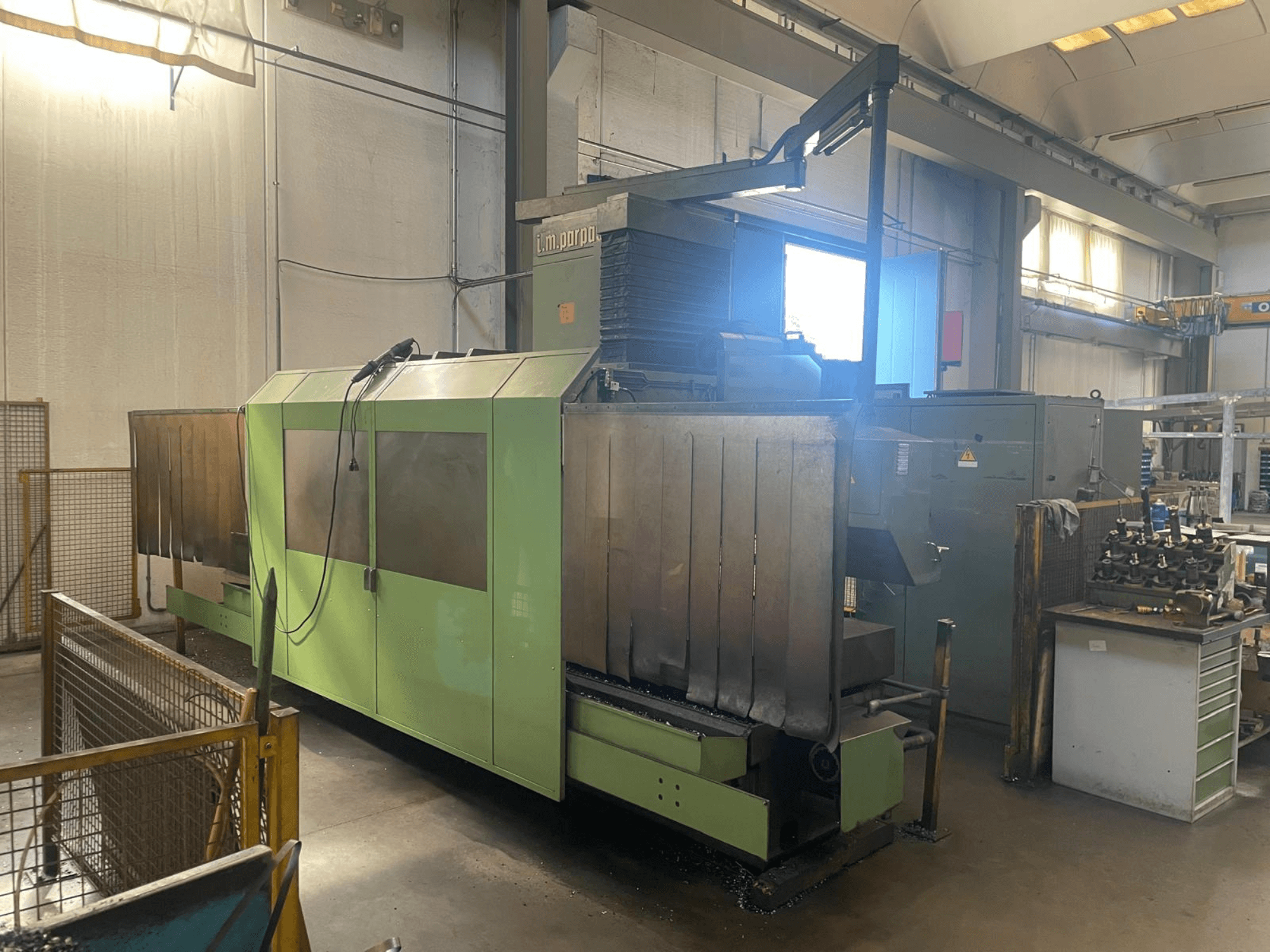 Macchina industriale verde con componenti metallici a vista, vista frontale; presenta pannello di controllo, avvisi di sicurezza e postazione di lavoro nelle vicinanze.