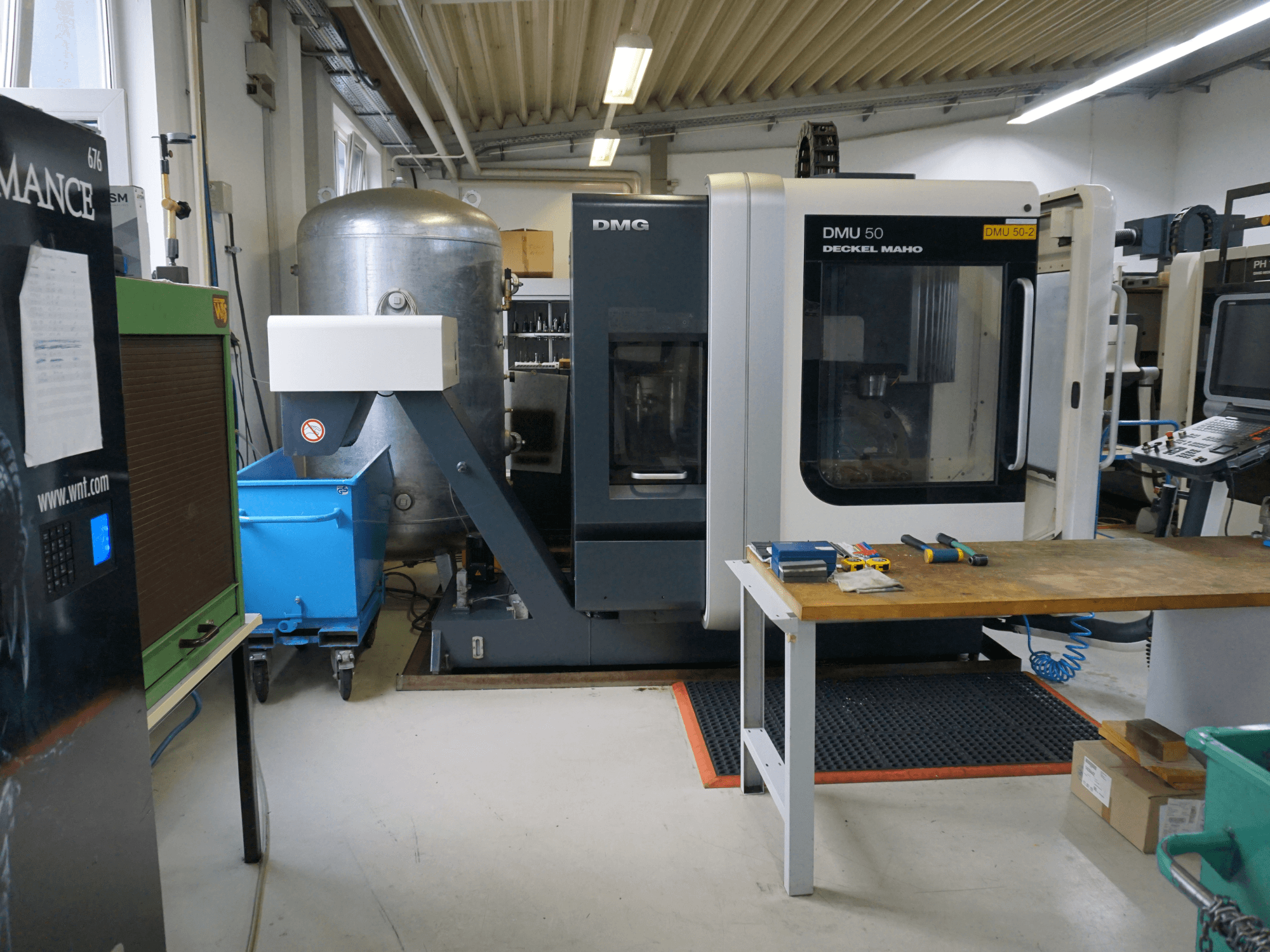 Fresatrice DMG DMU 50 in un'officina, vista frontale, con utensili su un tavolo di legno e un bidone blu a rotelle nelle vicinanze.