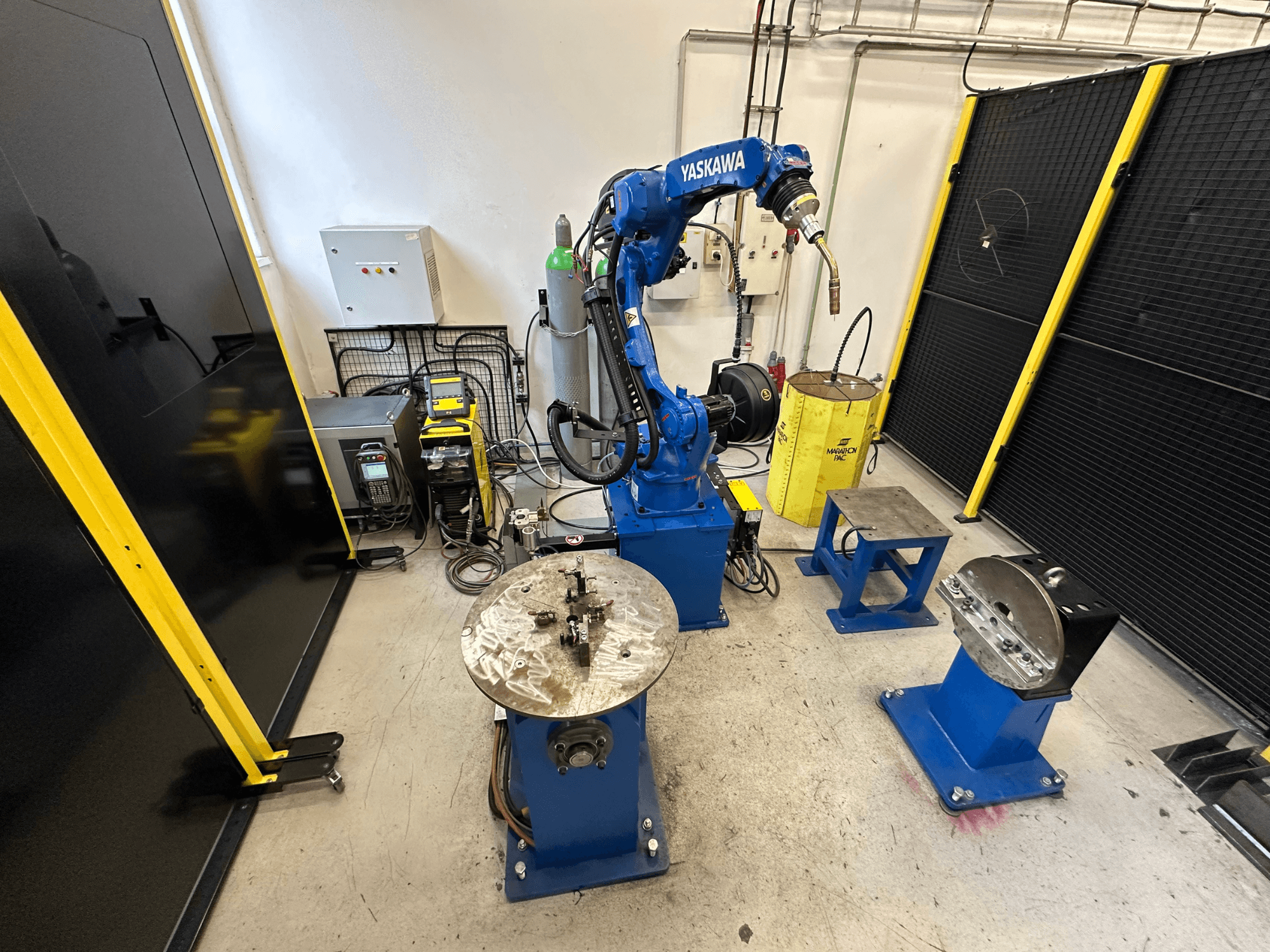Braccio robotico Yaskawa in blu con due tavoli da lavoro, attrezzature per la saldatura e barriere di sicurezza; vista frontale dell'area di lavoro industriale.