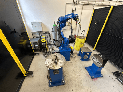 Braccio robotico Yaskawa in blu con due tavoli da lavoro, attrezzature per la saldatura e barriere di sicurezza; vista frontale dell'area di lavoro industriale.