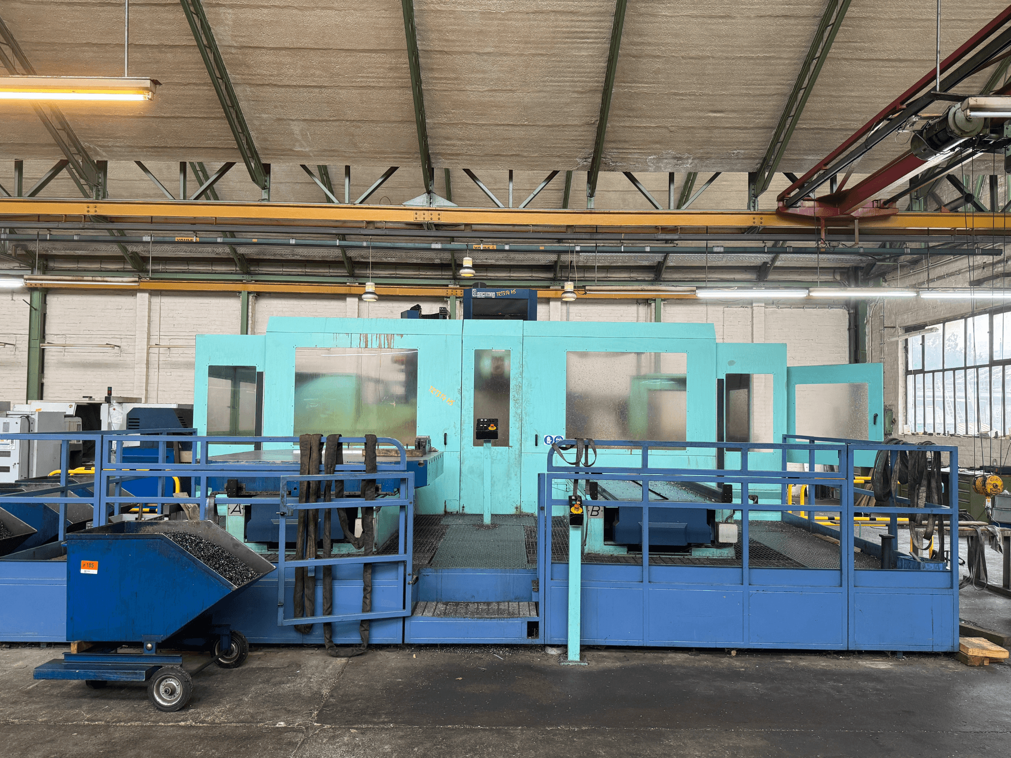 Macchina industriale a controllo numerico di colore blu con pannelli di protezione, vista frontale, dotata di magazzino utensili e carrello di raccolta rifiuti sul lato.