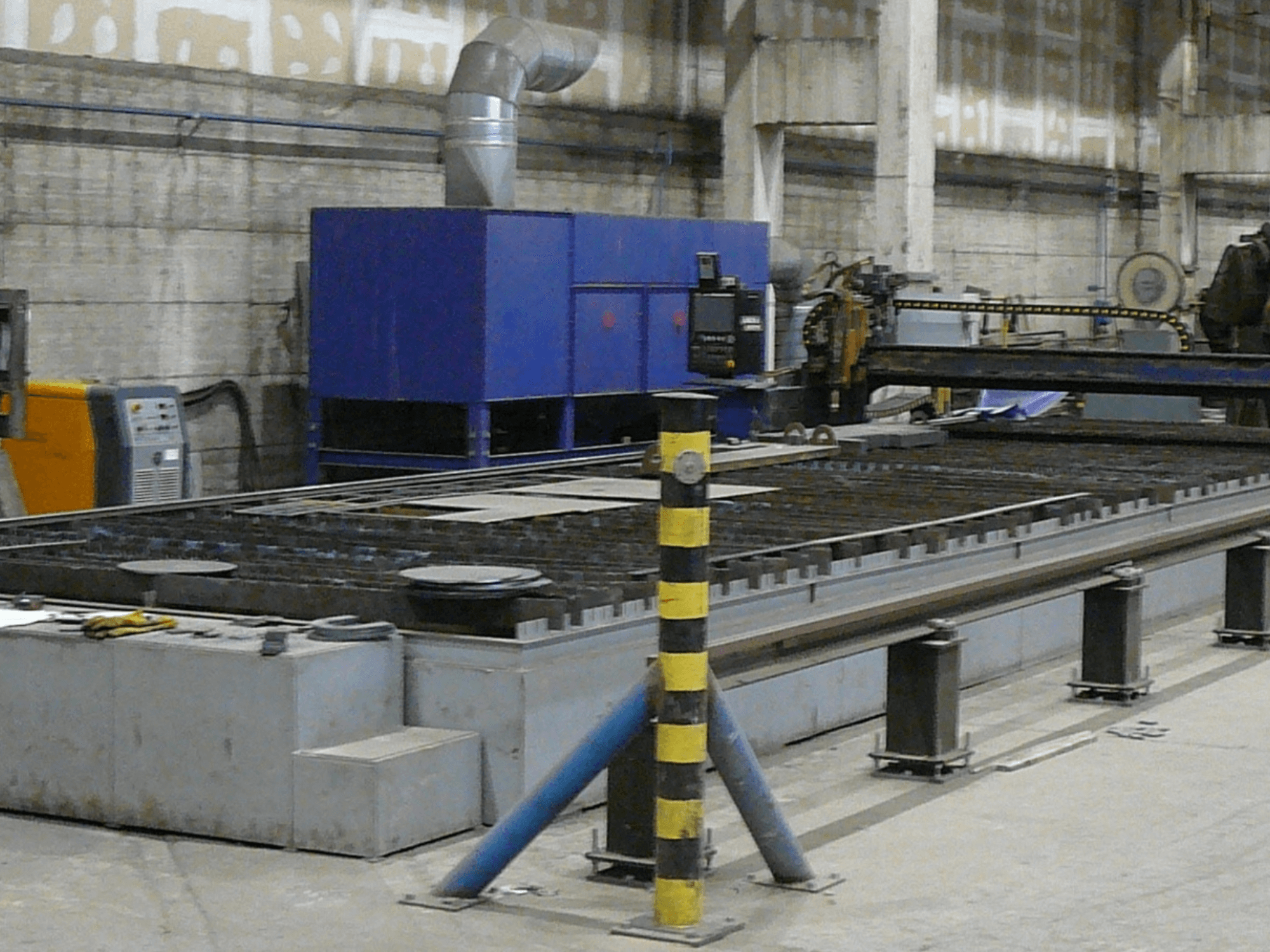 Configurazione di una macchina industriale per il taglio dei metalli, vista laterale, con quadro di comando blu, strisce di sicurezza e superficie di lavoro in metallo.