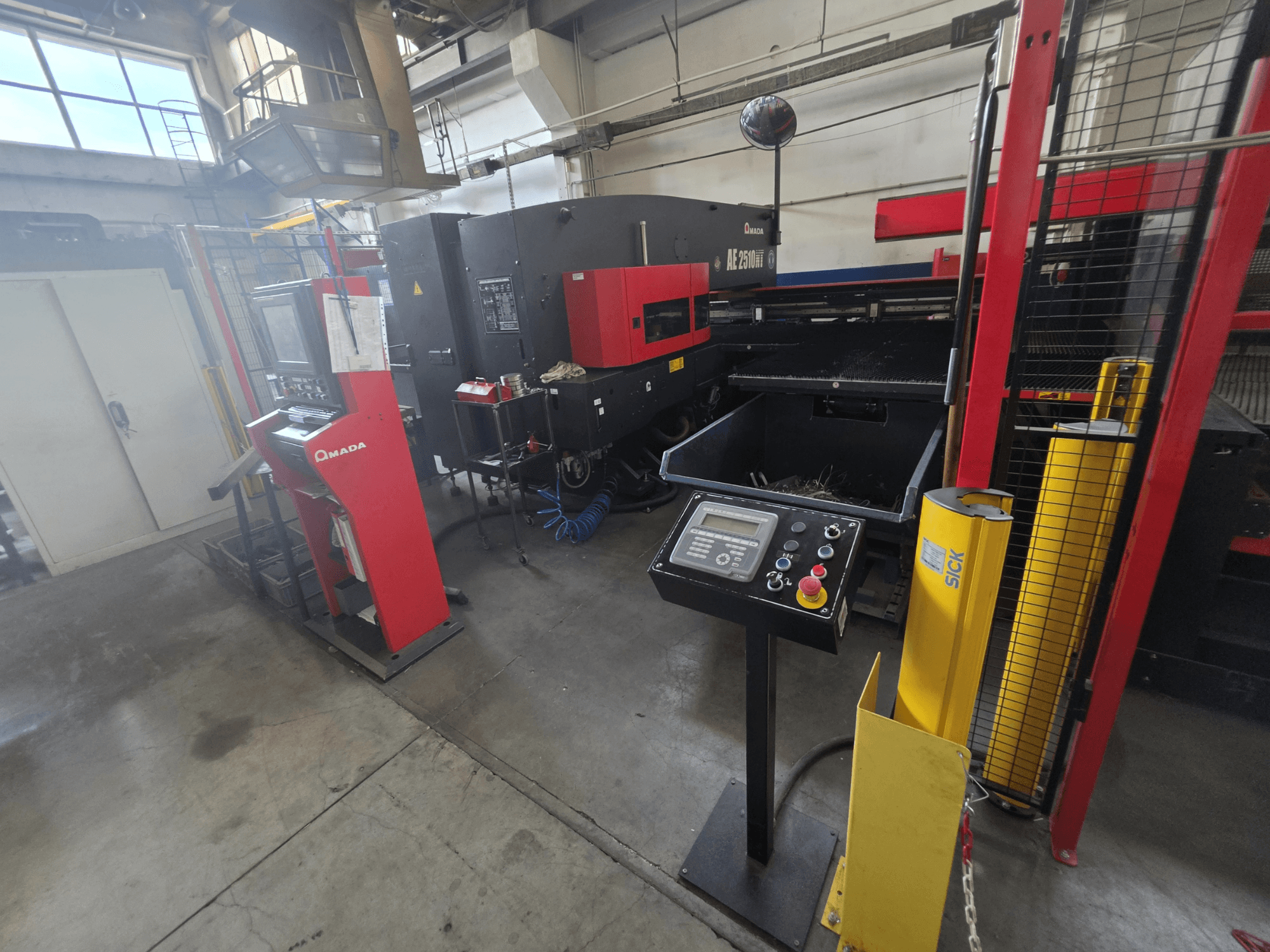 Punzonatrice CNC AMADA AE-2510NT, vista frontale con pannello di controllo, dispositivi di sicurezza e ambiente industriale circostante.