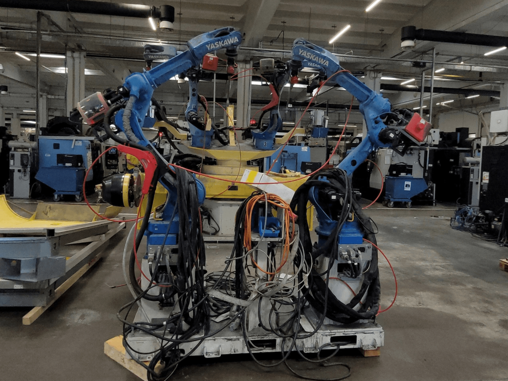 Cella di saldatura robotizzata Yaskawa RWC L1_OS 15330, vista frontale, con due robot blu dotati di cavi di notevole lunghezza e utensili di saldatura.