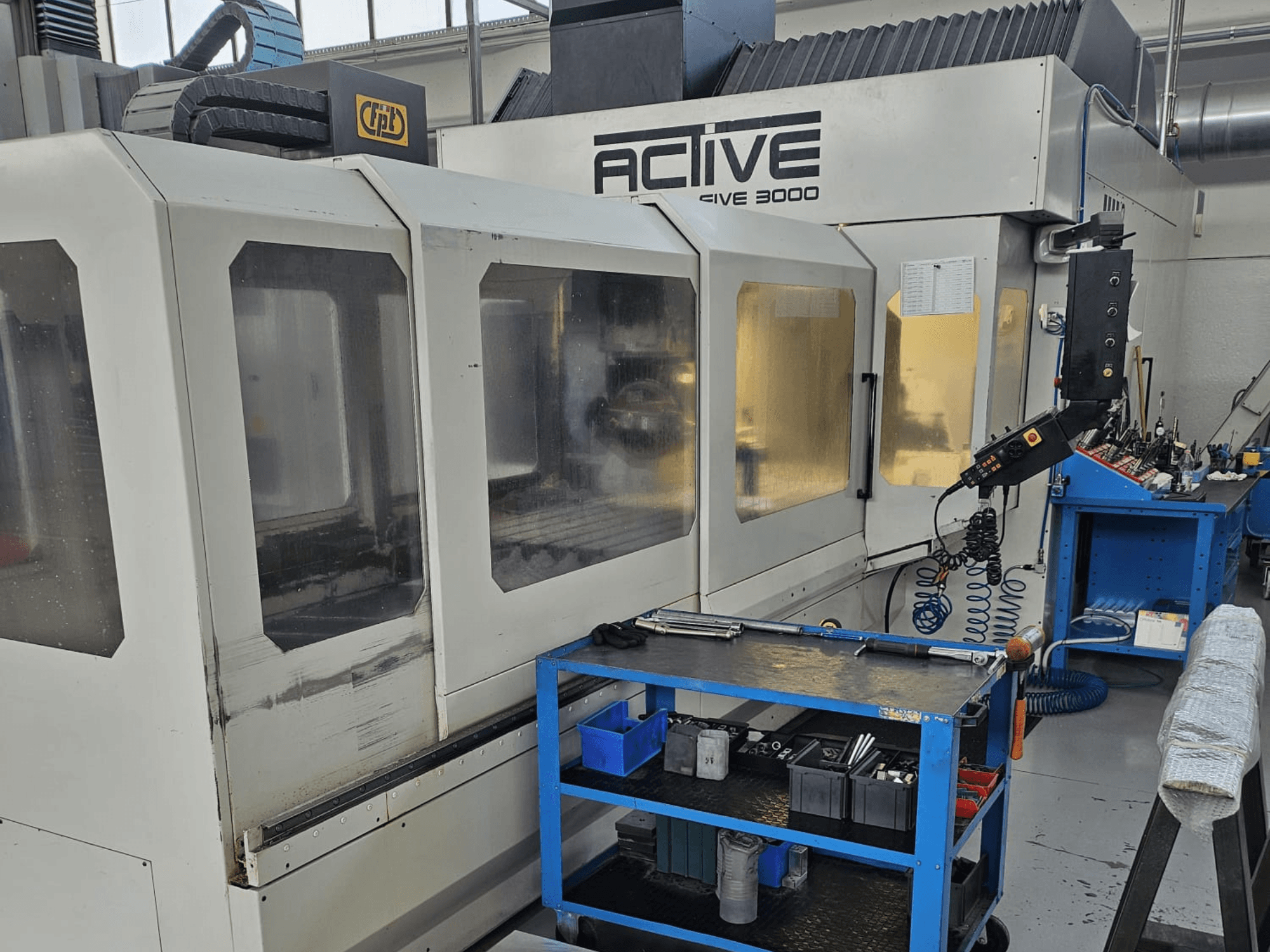 Macchina CNC Active 3000 in un'officina, vista frontale con utensili su un carrello blu, comandi visibili e area di lavoro interna.