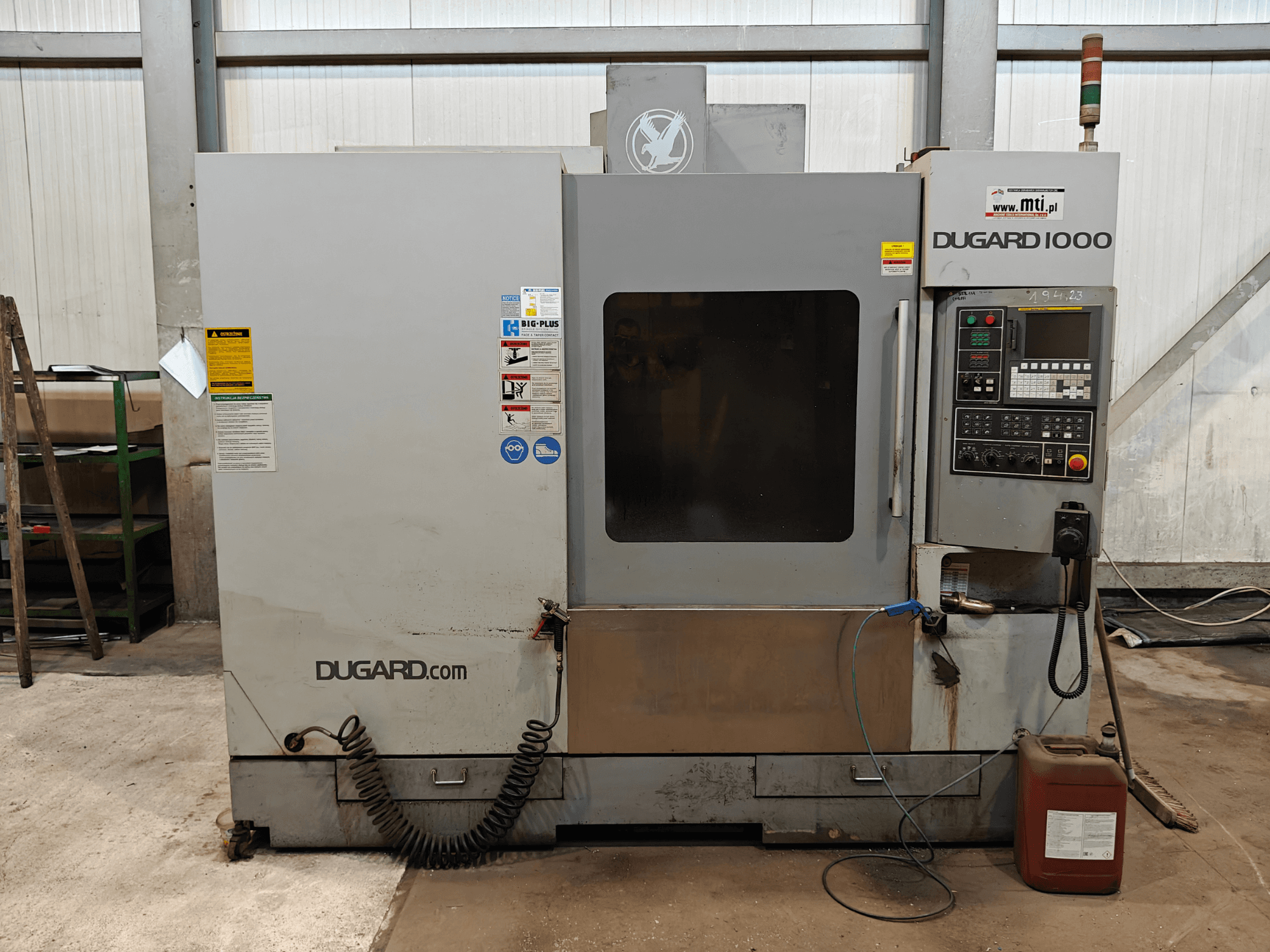 Macchina CNC Dugard 1000, vista frontale, con pannello di controllo, etichette di sicurezza e contenitore dell'olio, in un'officina.