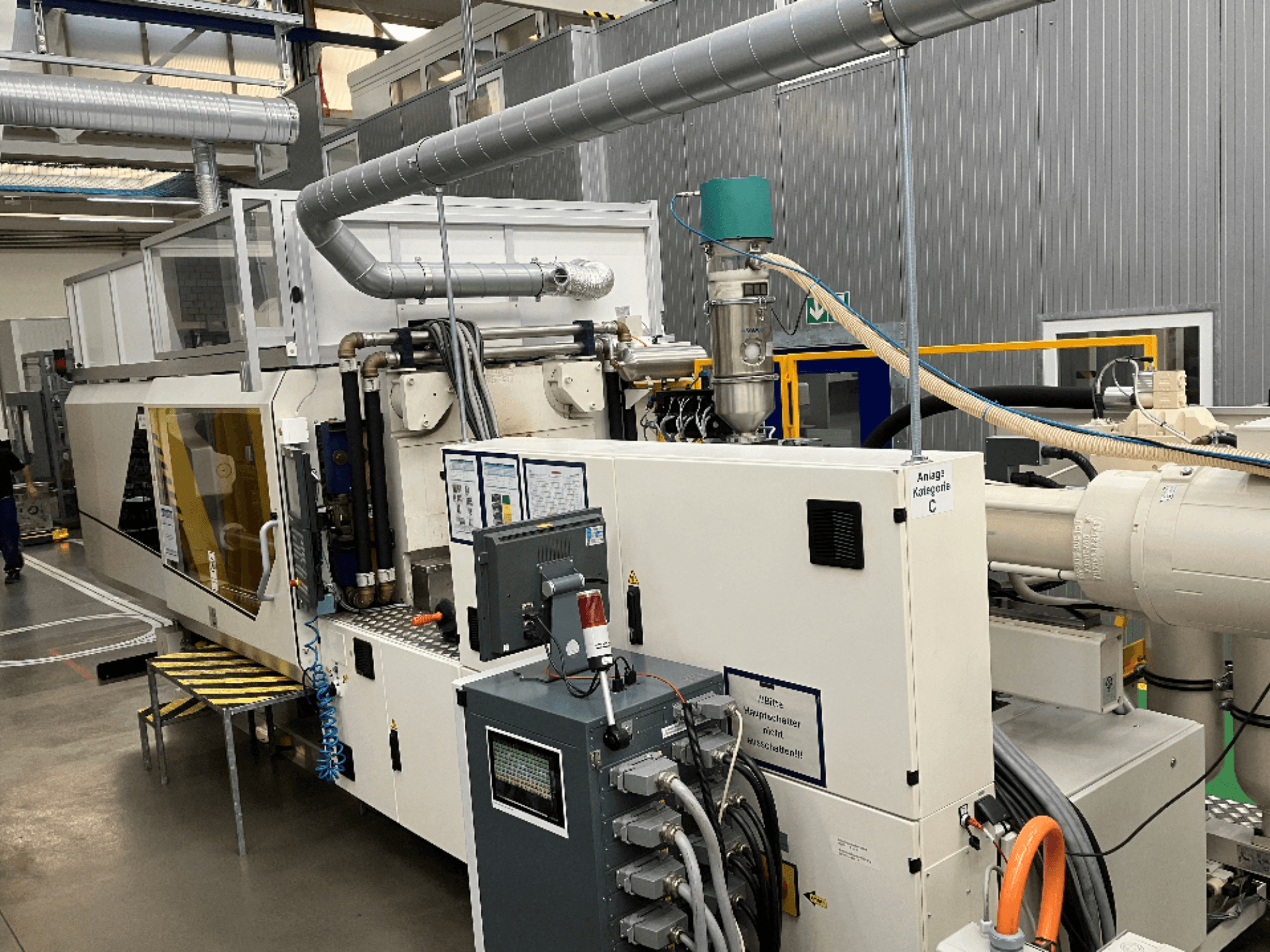 Macchina per lo stampaggio a iniezione, vista frontale, con etichetta del marchio, pannello di controllo e tubi flessibili visibili in un ambiente di fabbrica.