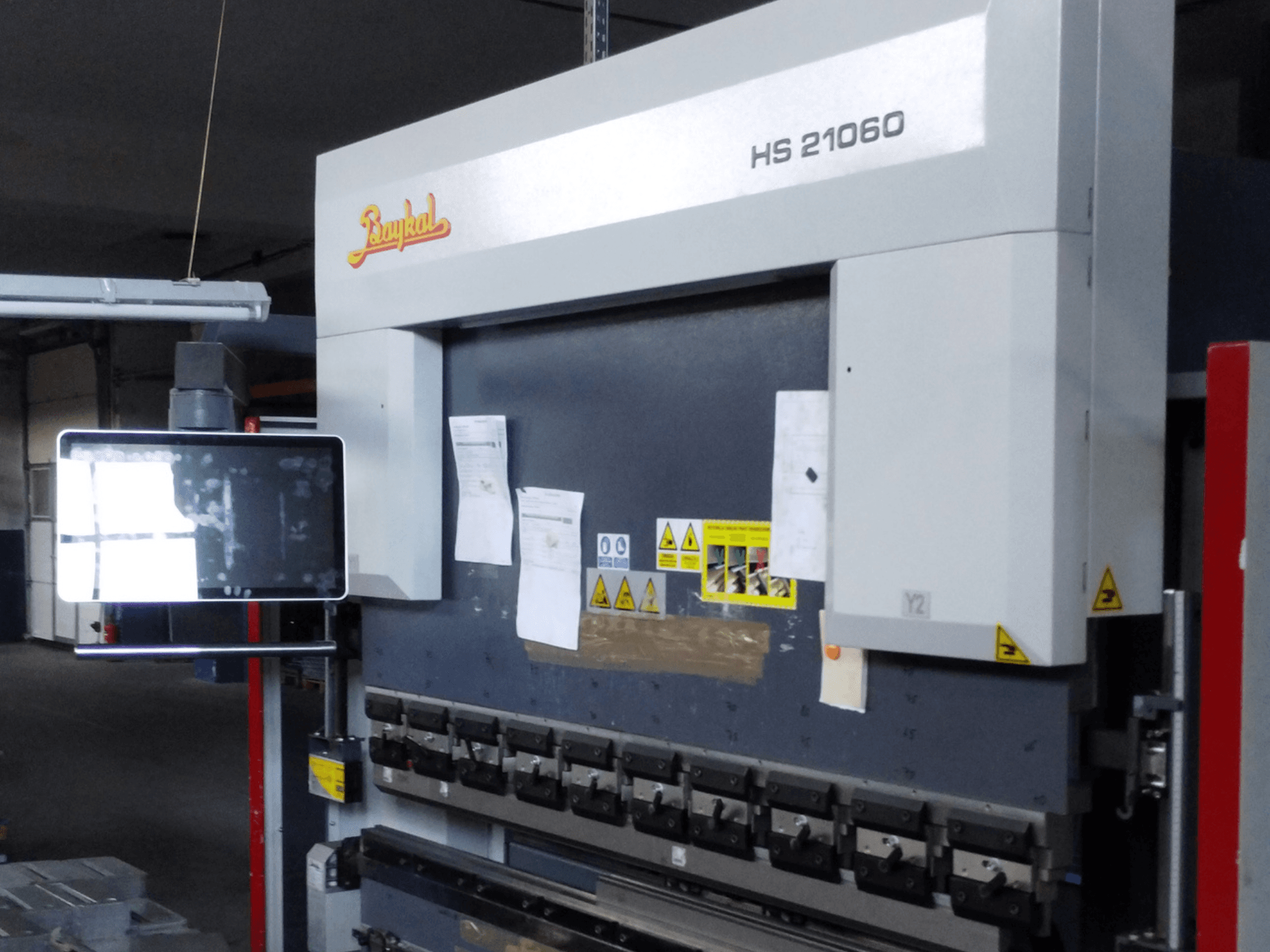 Vista frontale della pressa piegatrice Baykal APHS 21060, con display di controllo, avvertenze di sicurezza e utensili per la piegatura dei metalli.