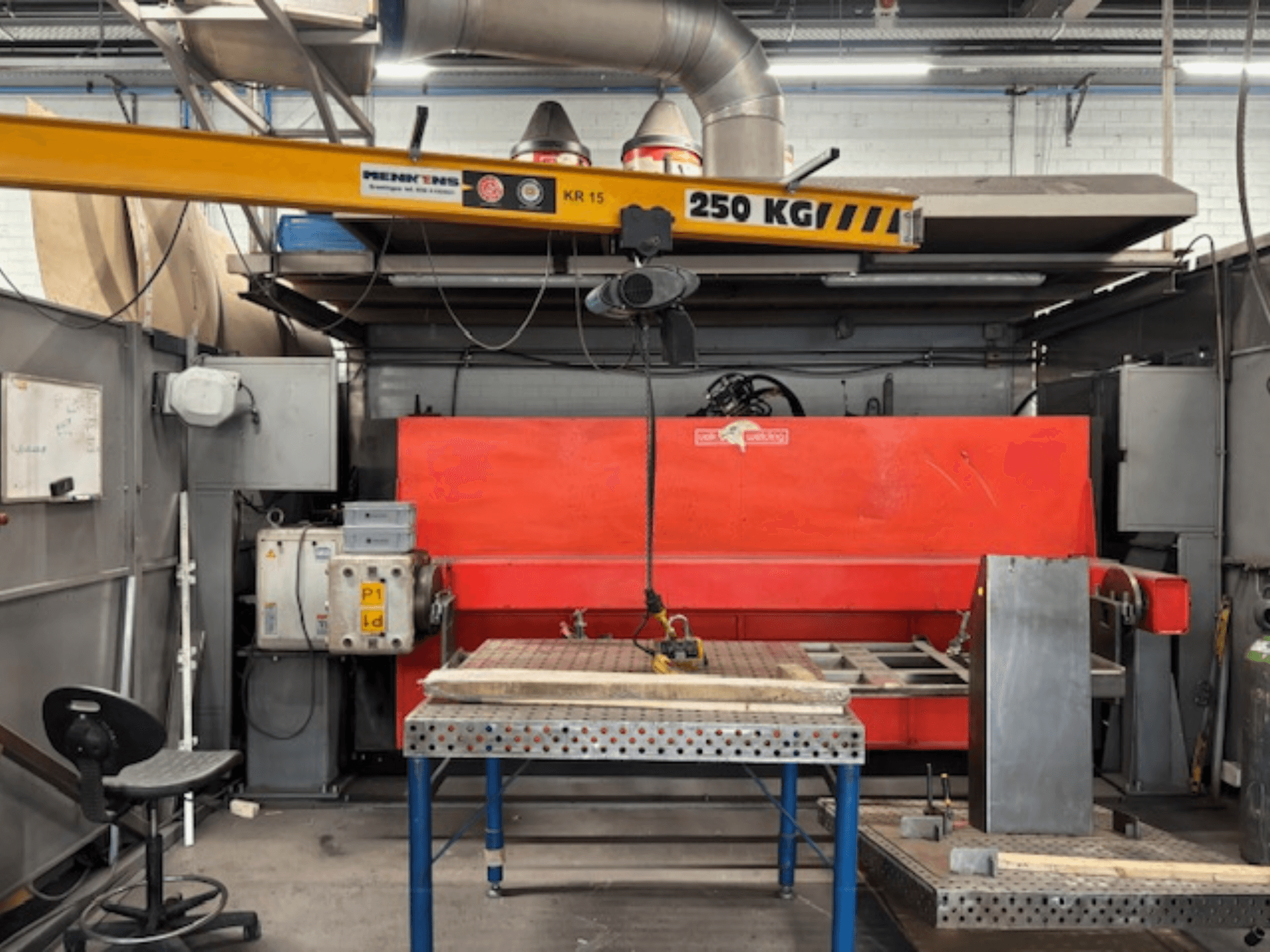 Officina industriale con una pressa idraulica rossa, un carroponte con la scritta "KR 15" e un tavolo da lavoro in metallo, prospettiva laterale.