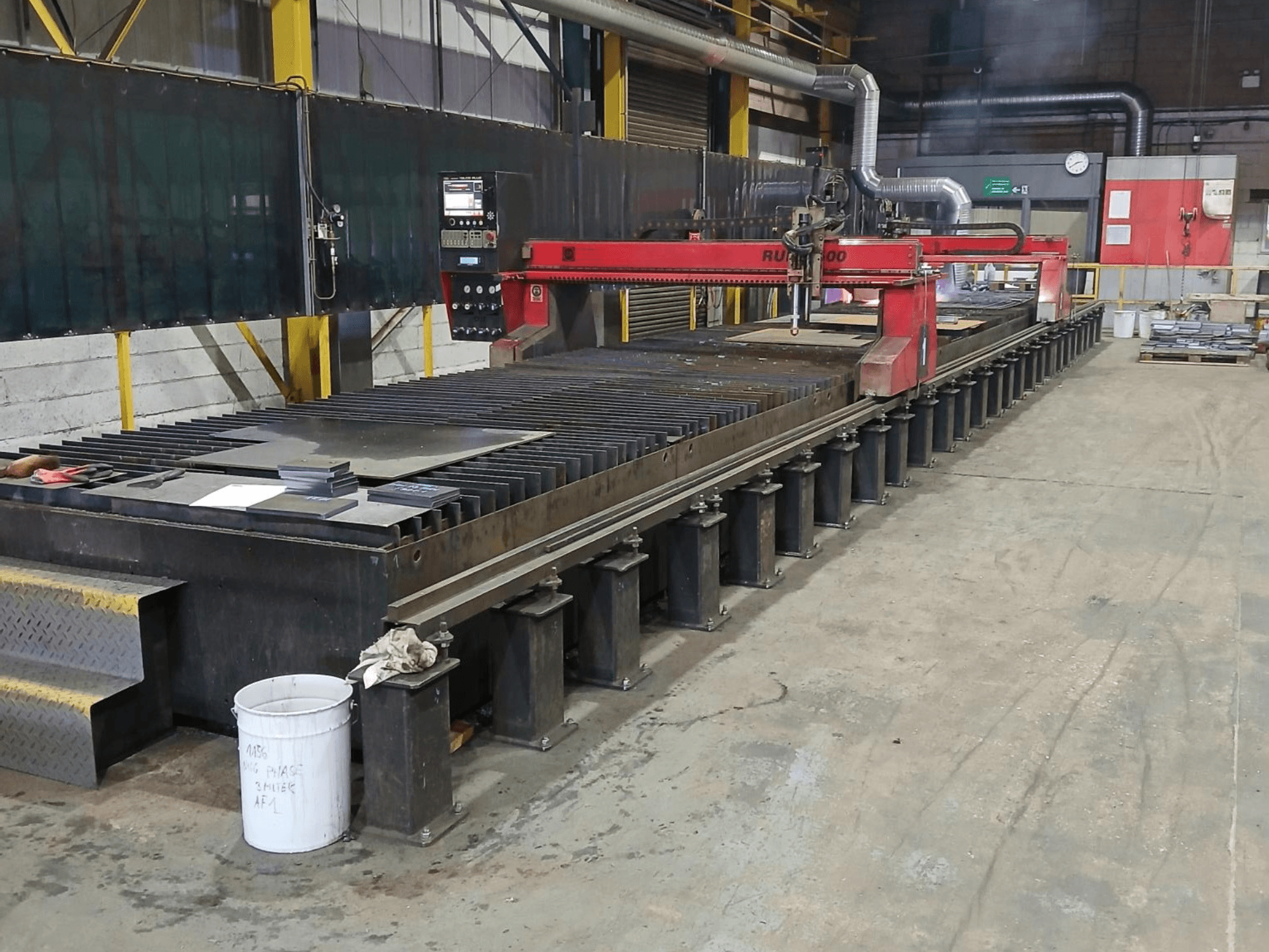 Macchina da taglio RUIDA 300 rossa, vista laterale, con lamiera su un tavolo d'acciaio, situata in uno spazio di lavoro industriale.