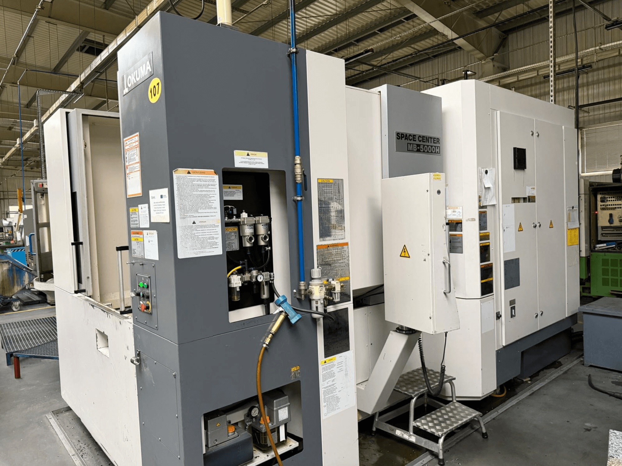 Centro di lavoro orizzontale Okuma MB 5000H, vista laterale che mostra il pannello di controllo e i componenti della macchina in un ambiente di fabbrica.