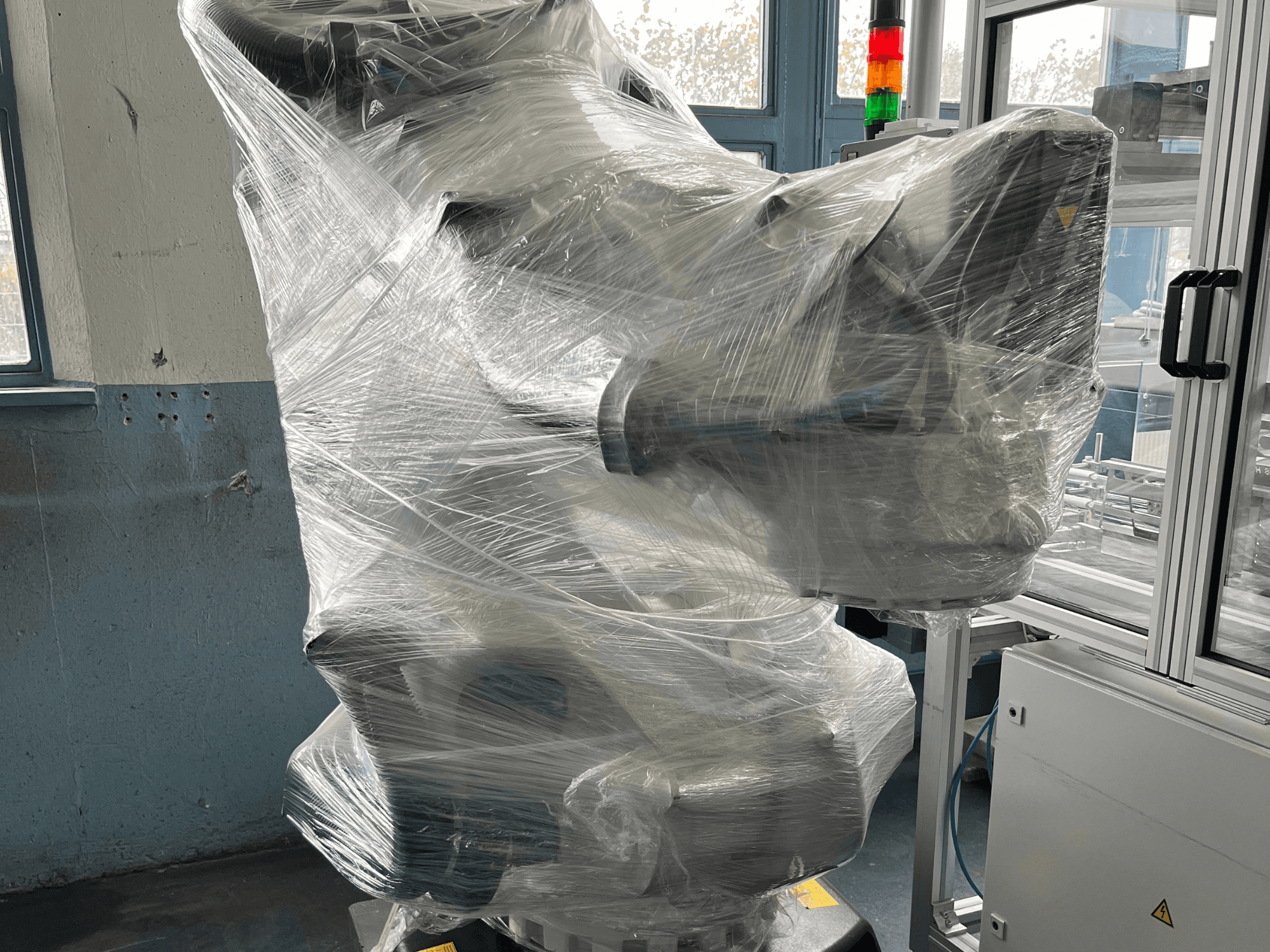 Braccio robotico industriale avvolto in plastica, vista frontale. Include unità di controllo e spia luminosa. Marchio non visibile.