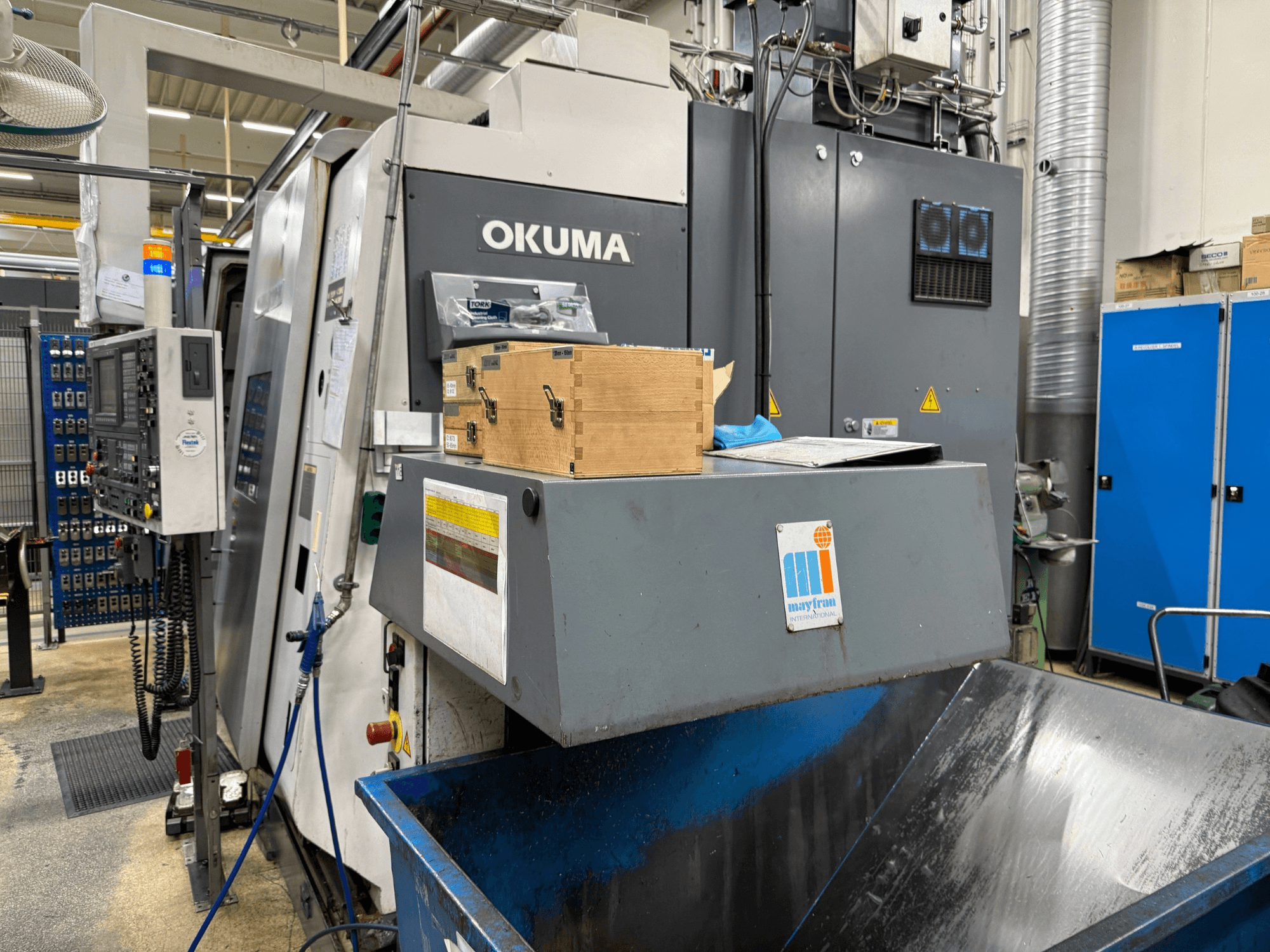 Macchina CNC Okuma in fabbrica, vista frontale; presenta il pannello di controllo, le scatole di legno e il cestino blu con il logo Mayfran.