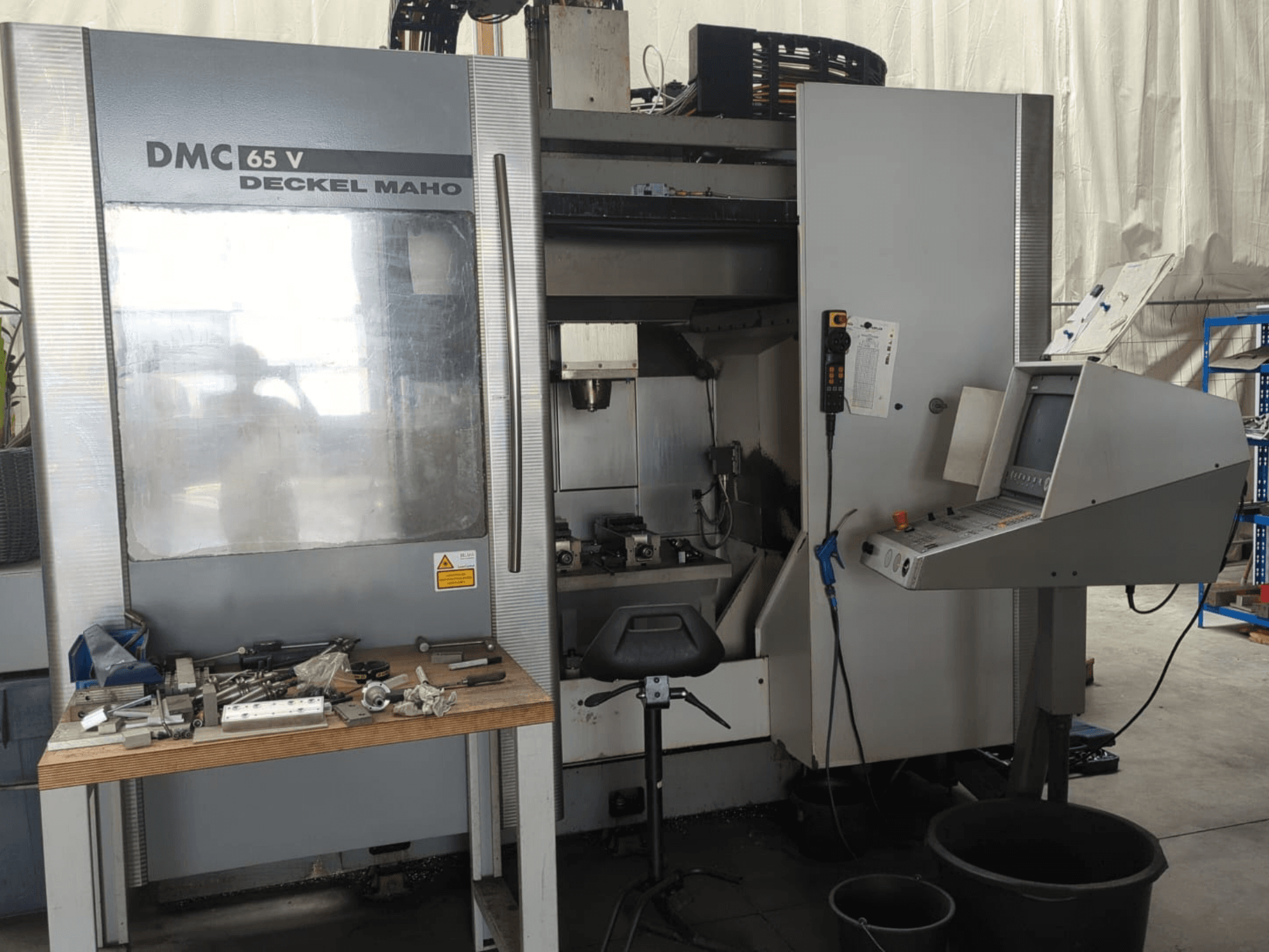 Vista frontale di un centro di lavorazione verticale DECKEL MAHO DMC 65 V con pannello di controllo, utensili sul tavolo e configurazione dell'area di lavoro.