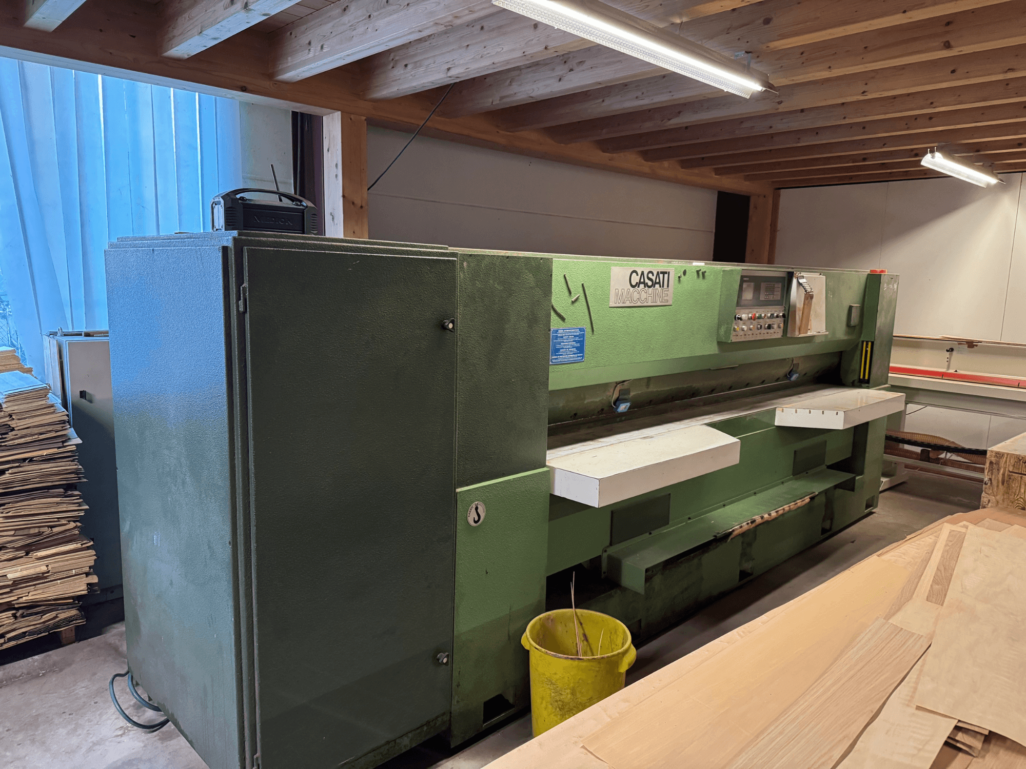 Macchina per la lavorazione del legno CASATI verde in un'officina, vista frontale, con pannello di controllo e superfici di lavoro bianche, circondata da cataste di legna.