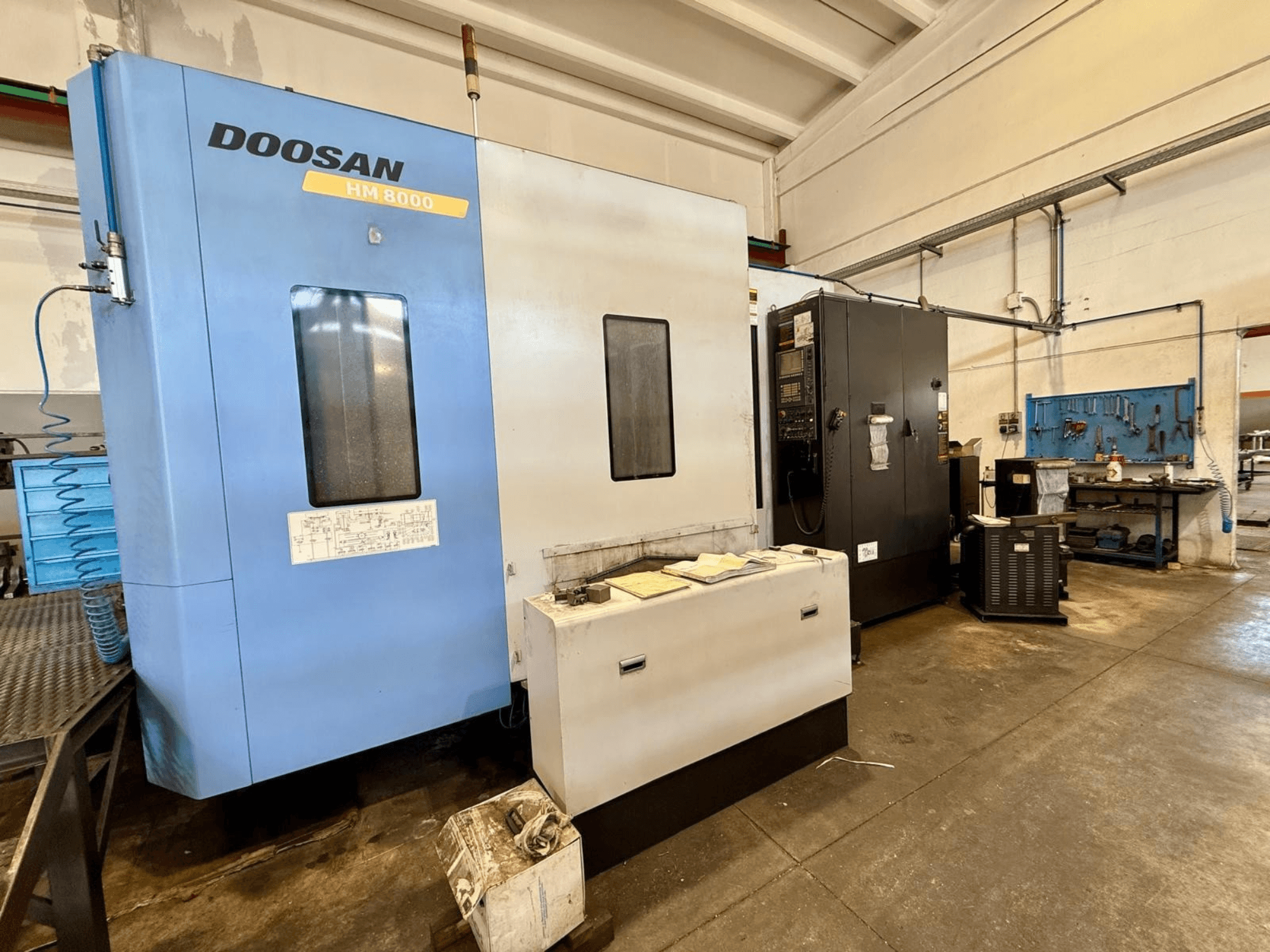Centro di lavorazione orizzontale DOOSAN HM 8000 di colore blu, vista frontale, con pannello di controllo e attrezzature dell'officina nelle vicinanze.