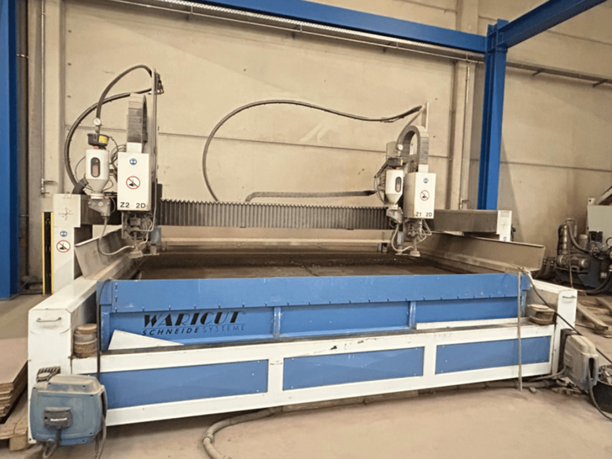Macchina da taglio a getto d'acqua di Waricut, vista frontale; presenta due teste di taglio e una superficie di lavoro blu in un'officina.