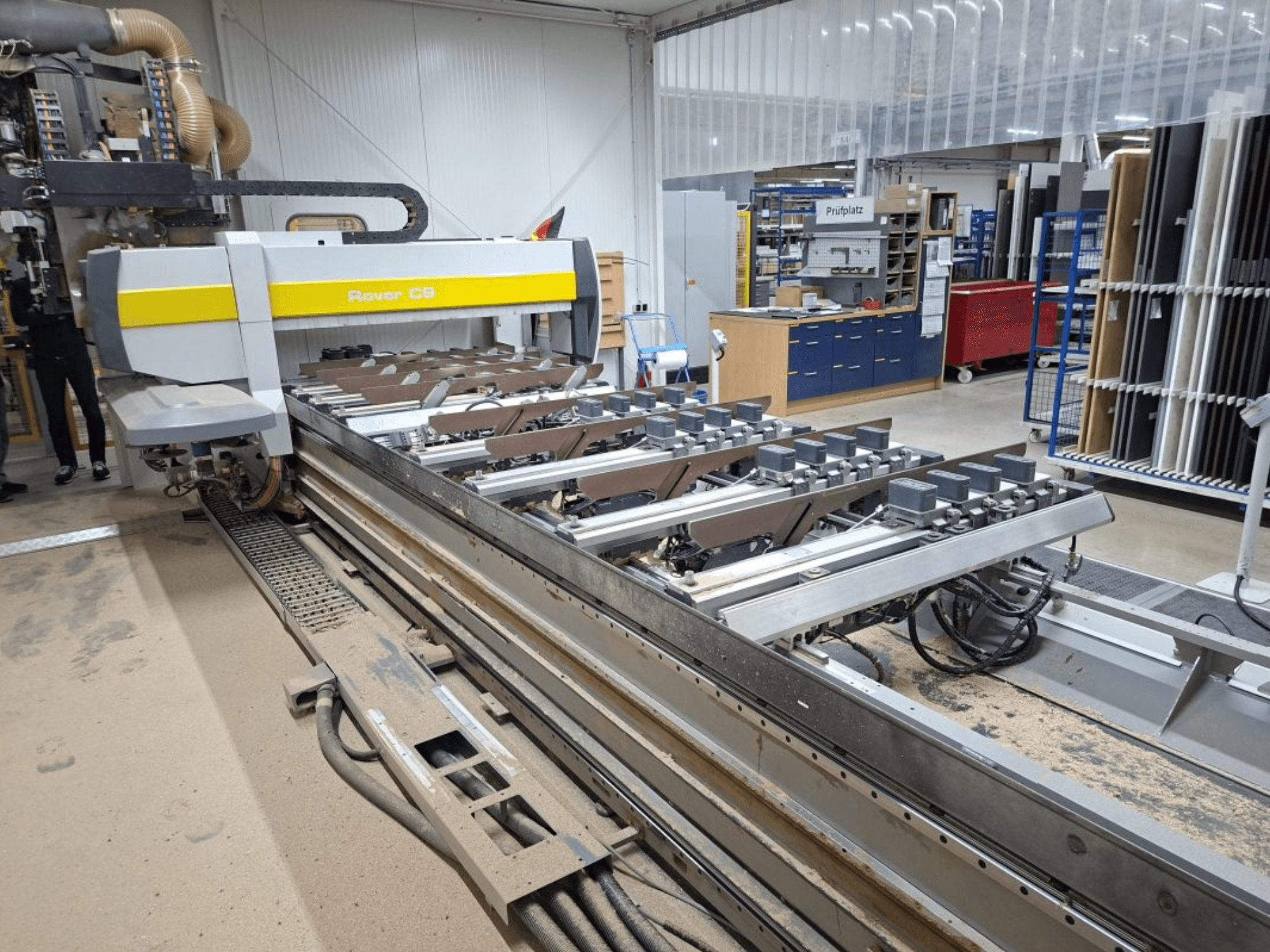 Macchina CNC Rover C9 in un'officina; la vista frontale mostra gli utensili da taglio e il sistema di trasporto, con accumulo di polvere sul pavimento.