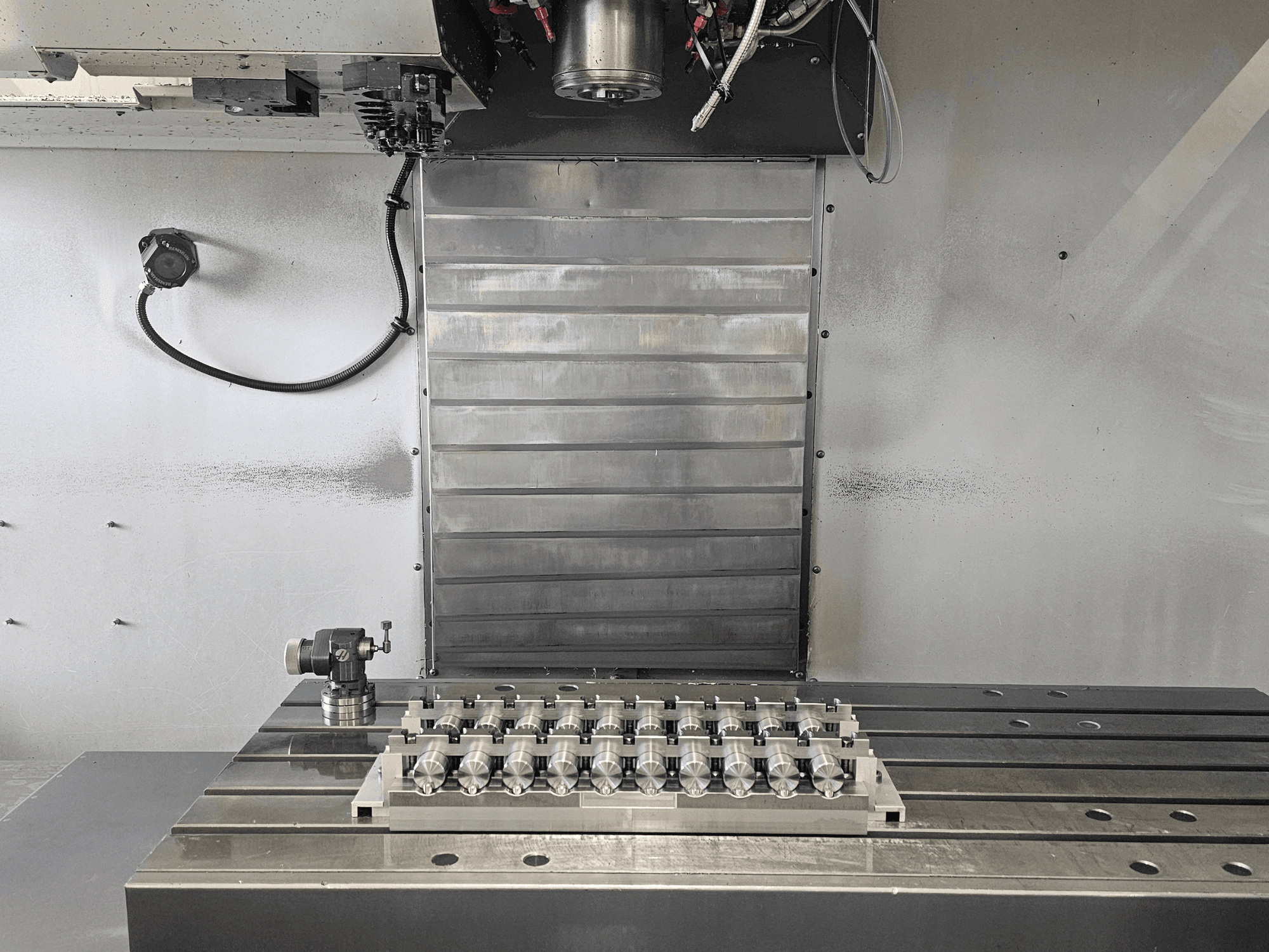 Vista frontale dell'area di lavoro di una fresatrice CNC con un pezzo lavorato di precisione su una tavola metallica e meccanismi di controllo.