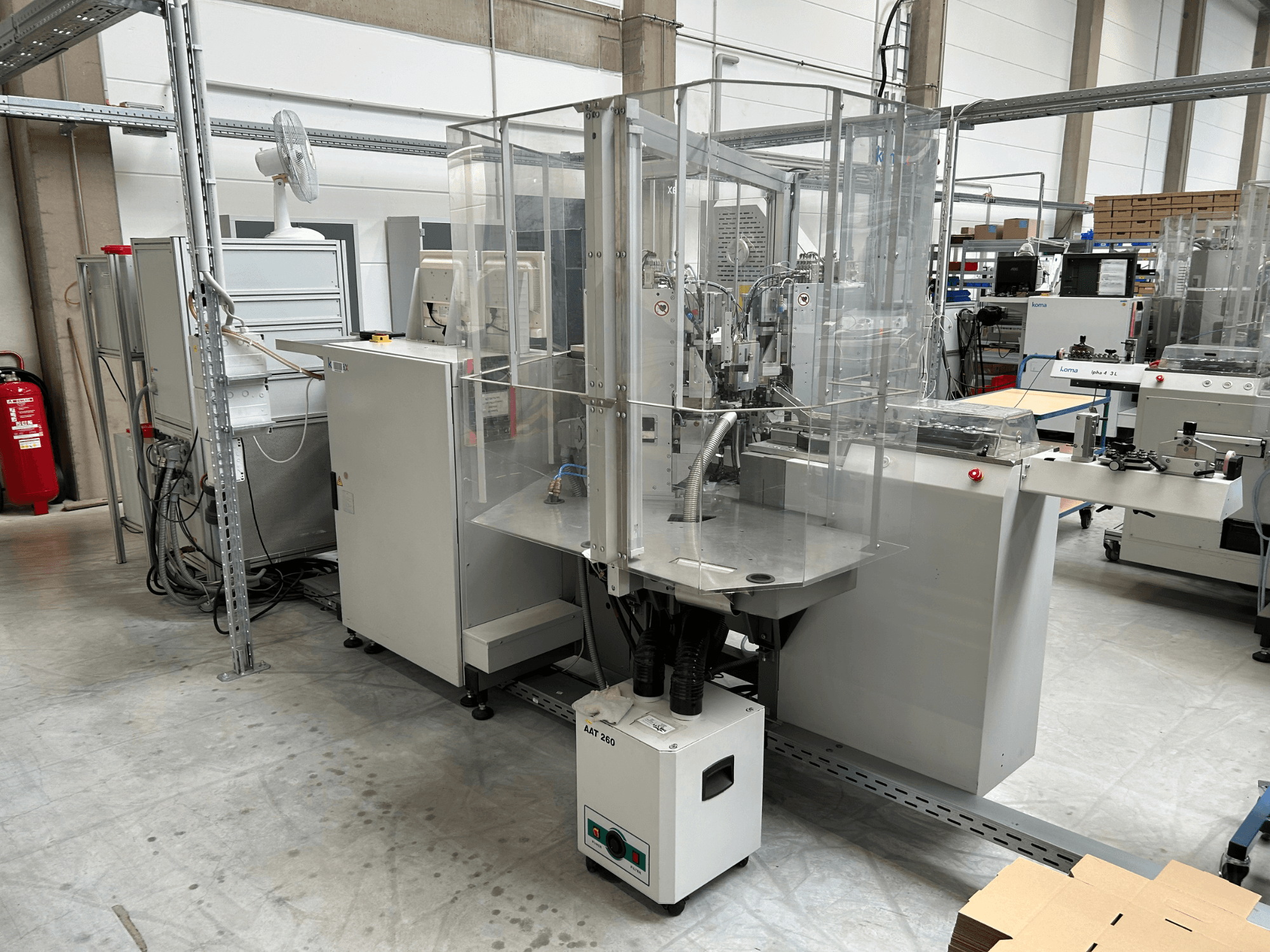 Apparecchiatura di automazione KOMAX Alpha 433 L in un ambiente industriale. È dotata di una cabina di sicurezza trasparente e di vari pannelli di controllo.