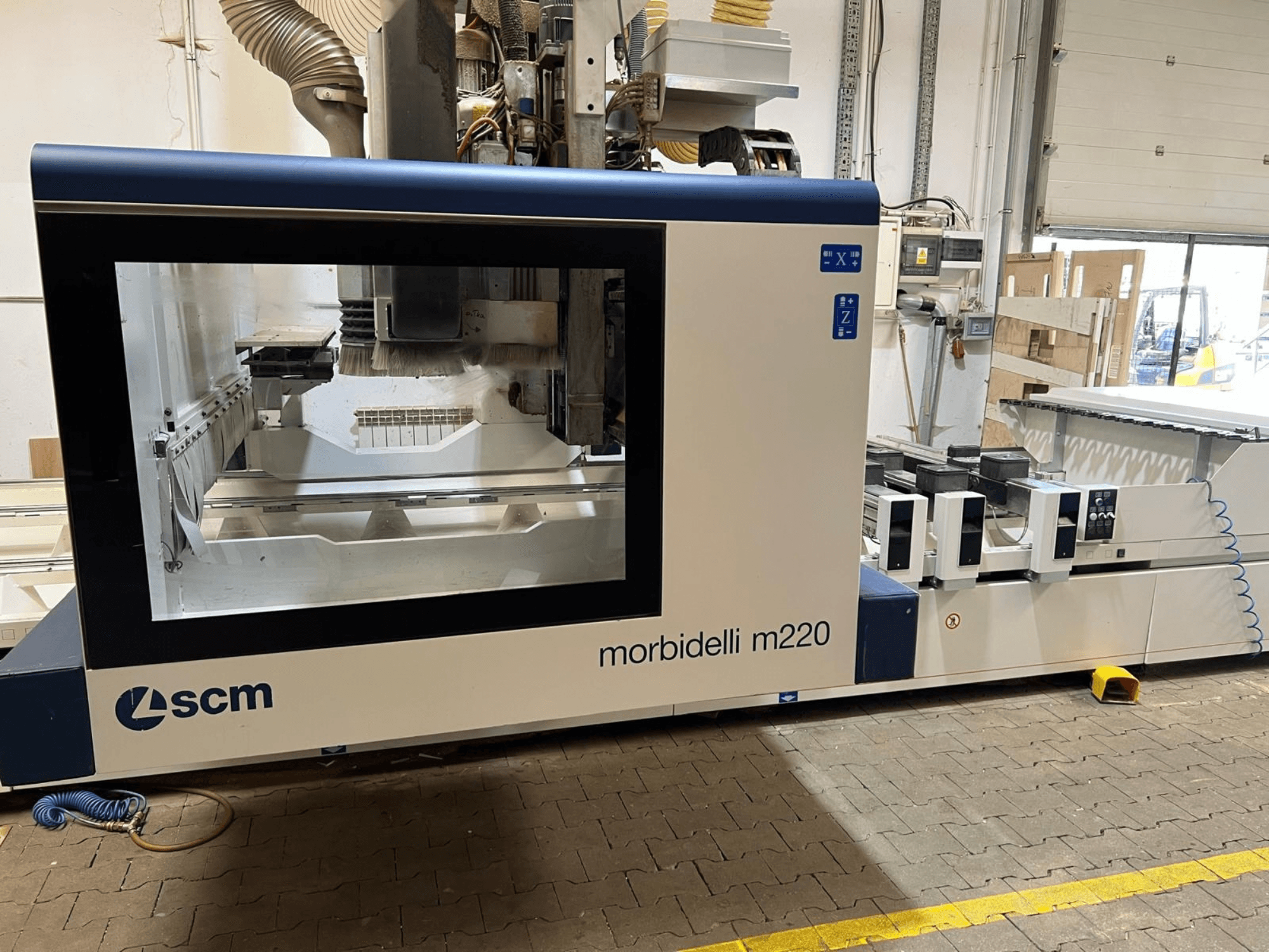 Vista frontale del centro di lavorazione CNC SCM Morbidelli M220, con in primo piano un'ampia finestra, il pannello di controllo e l'area di lavoro.