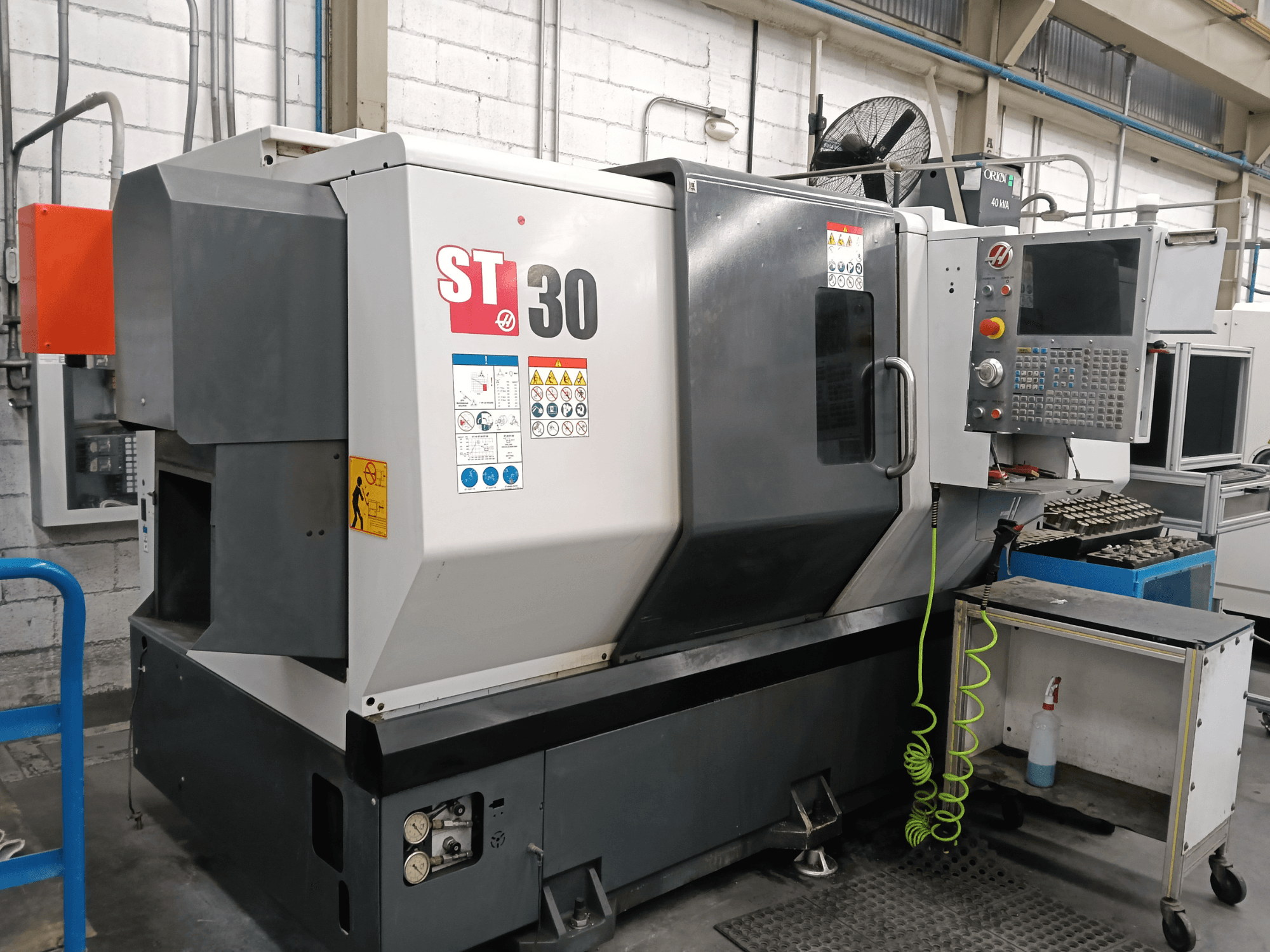 Testo alternativo: Vista frontale del tornio orizzontale HAAS ST-30 con pannello di controllo, etichette di sicurezza e utensili su un carrello nelle vicinanze.