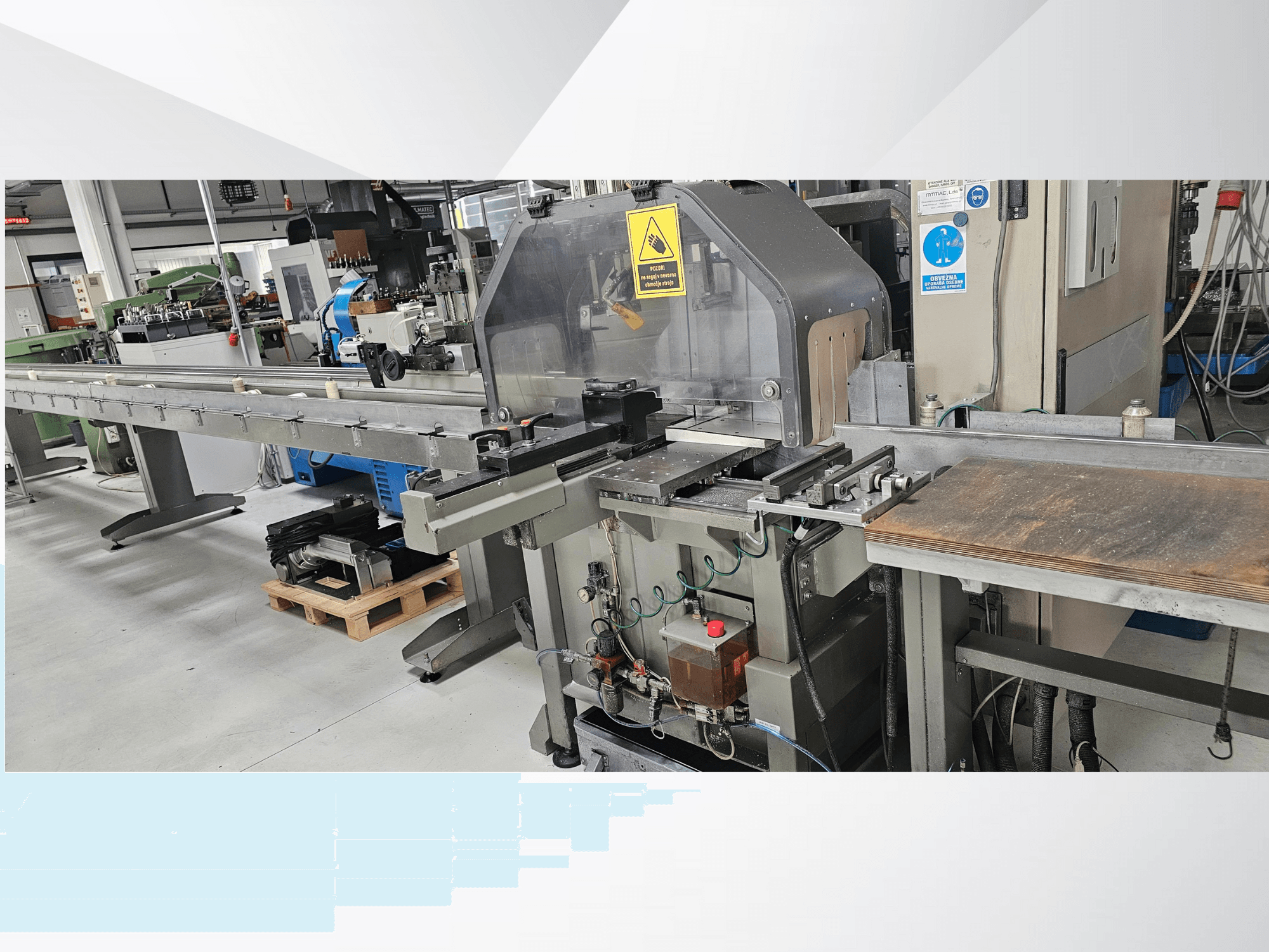 Macchina da taglio industriale vista di lato, con un lungo nastro trasportatore, un pannello di controllo e avvisi di sicurezza.