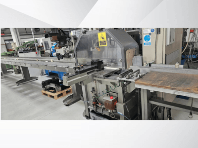 Macchina da taglio industriale vista di lato, con un lungo nastro trasportatore, un pannello di controllo e avvisi di sicurezza.