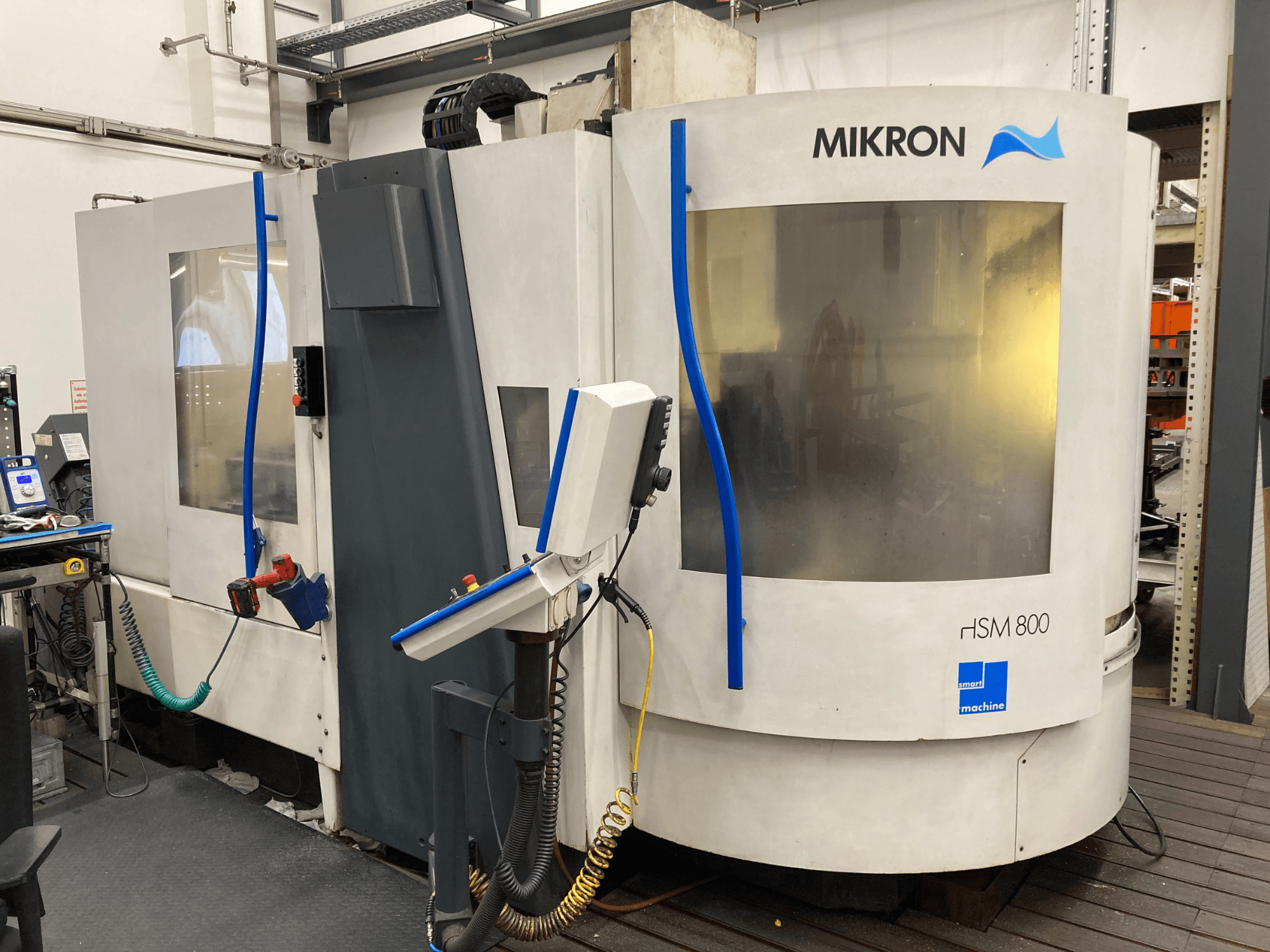 Mikron HSM 800, vista laterale di un centro di lavoro CNC con pannello di controllo e portautensili, tubi e cavi visibili.