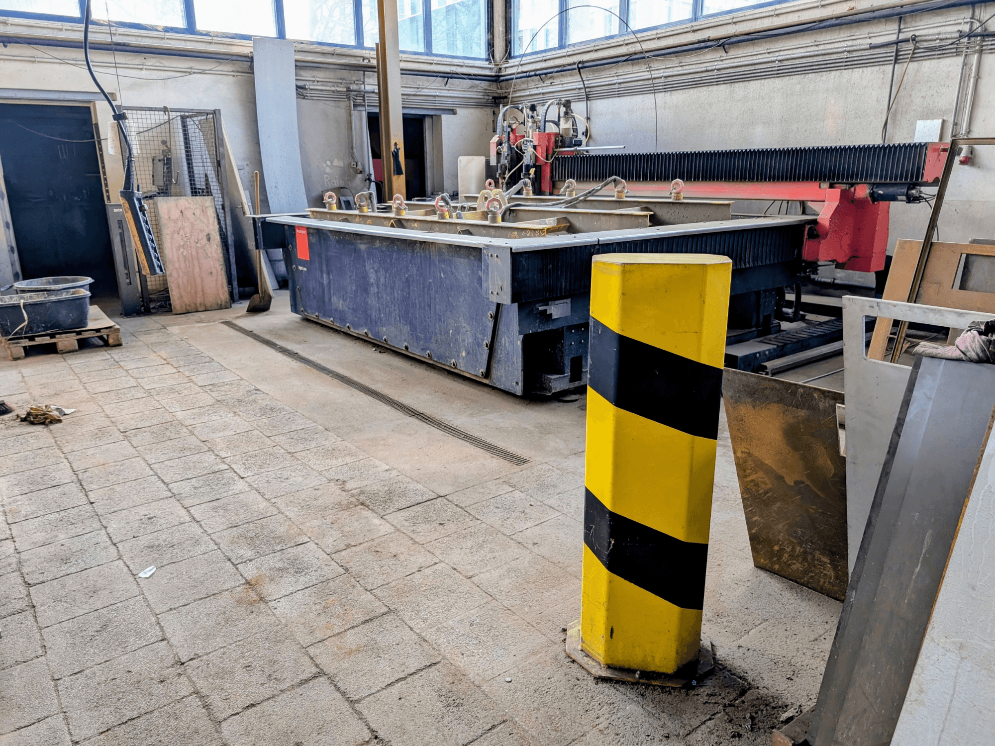 Macchina da taglio a getto d'acqua Bystronic Byjet L 2030 in un'officina, vista laterale, con palo di sicurezza giallo e materiali circostanti.