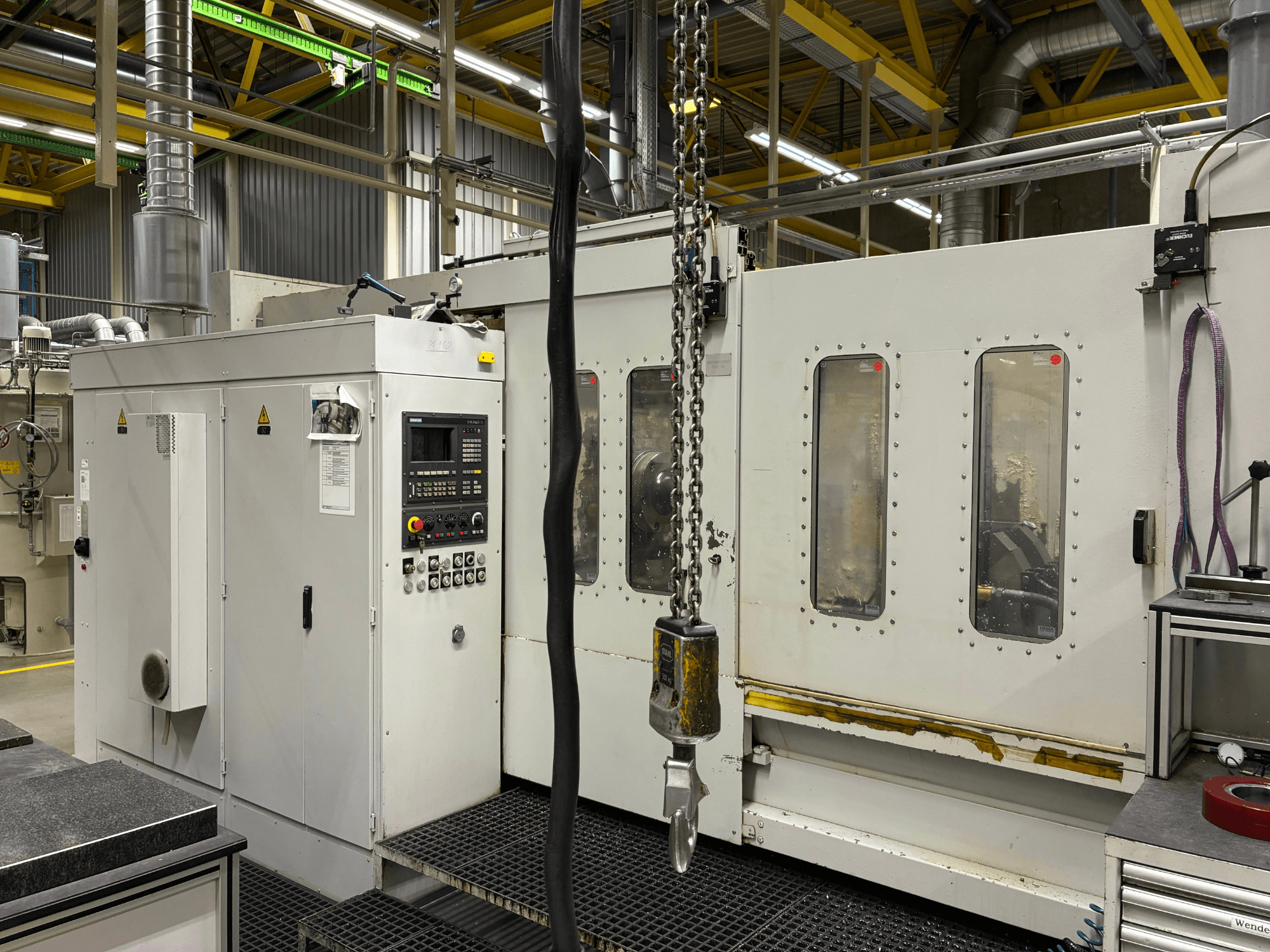 Macchina CNC industriale con pannello di controllo, vista laterale; presenta finestre, catene e componenti metallici in un ambiente di fabbrica.